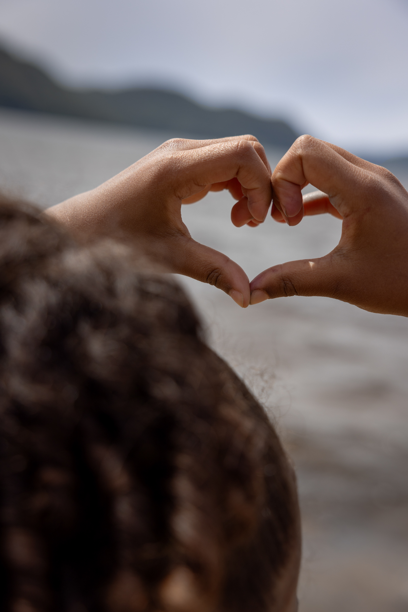 Foto: Barnehender som holder opp begge hendene og danner et hjerte med fingrene. Gjennom hjerte sees strand og sjø.