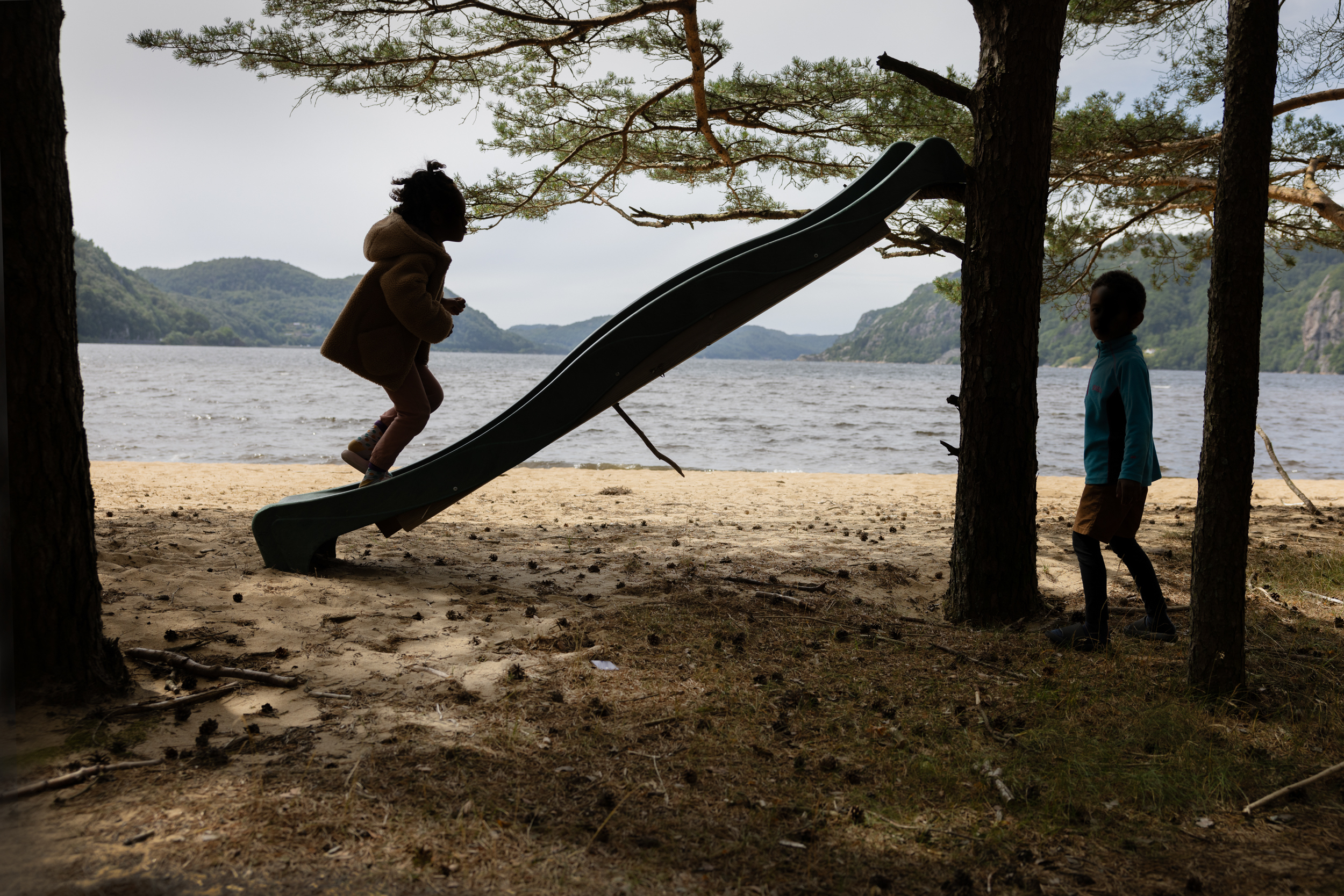Foto: To barn i skogen som leker på/ved en huske. I bakgrunnen sees sjøen.