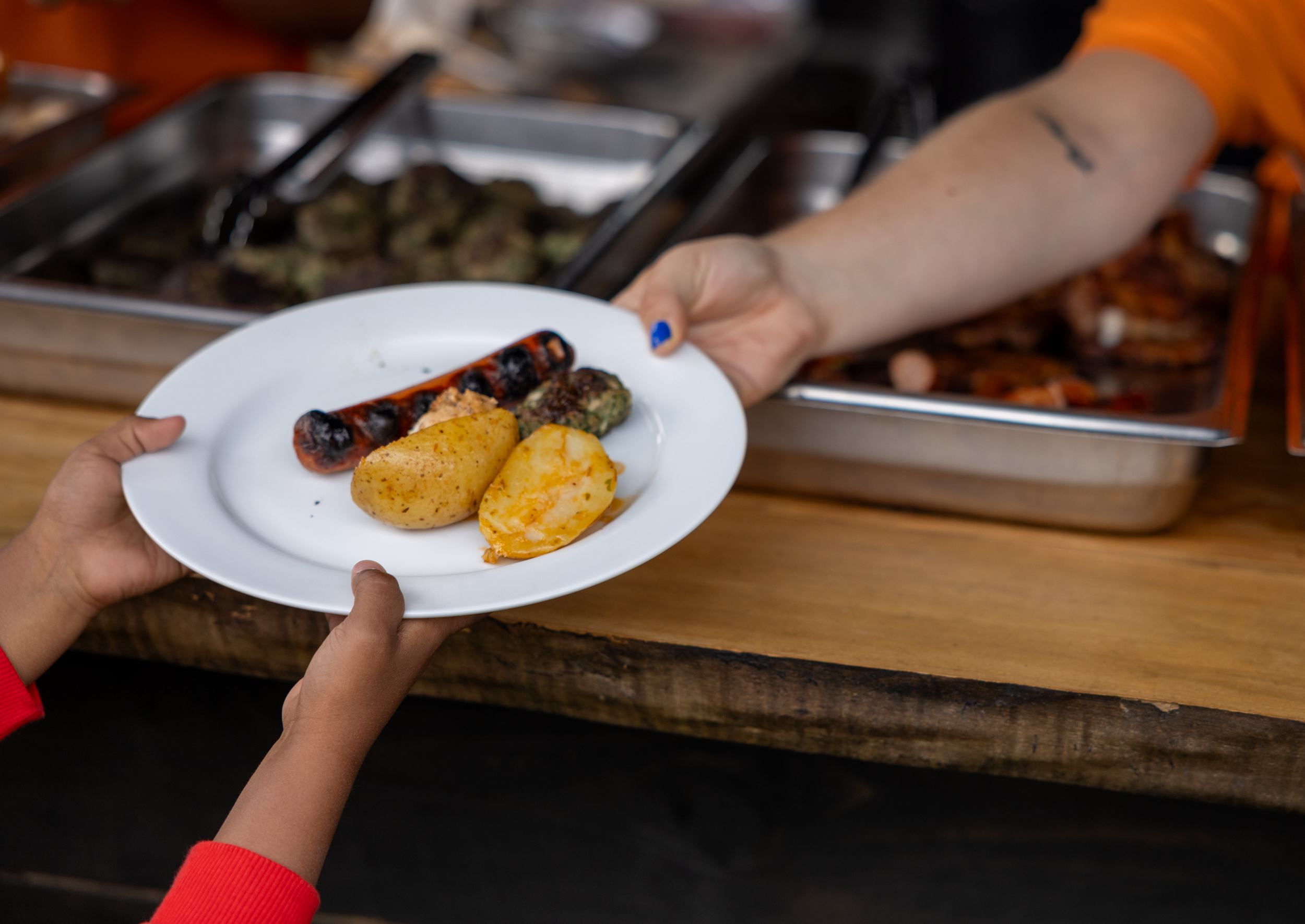 To foto: Det første viser en dame fra Sør-Amerika og en blond jente med fletter, si en grillhytte, om serverer kylling og annet kjøtt til en dame og to jenter med ryggen til kamera. Det andre bildet viser en voksen hånd som gir en tallerken med poteter og to pølser, og to barnehender som mottar.