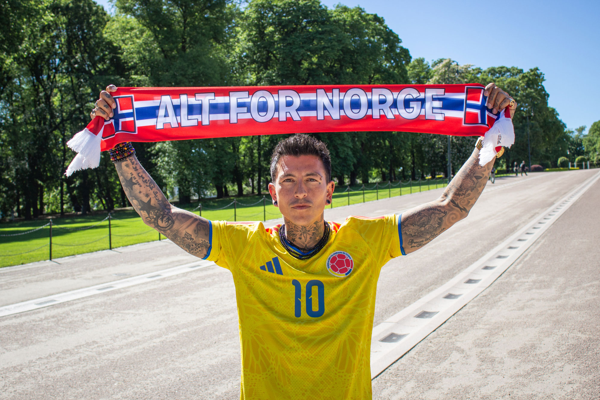 Foto: Mann i 30-årene med mørkt hår og mange tatoveringer holder opp et flagg der det står "Alt for Norge" i rødt, hvitt og blått. Han har på seg en gul fotballtrøye med blå og rød detaljer der det står tallet 10 foran.