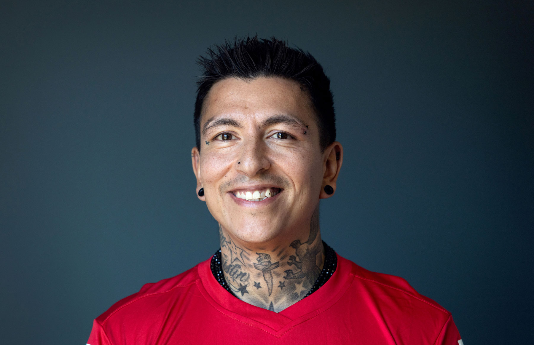 Foto: Mann med svart hår og tatoveringer smiler. Han har på seg en rød drakt.
