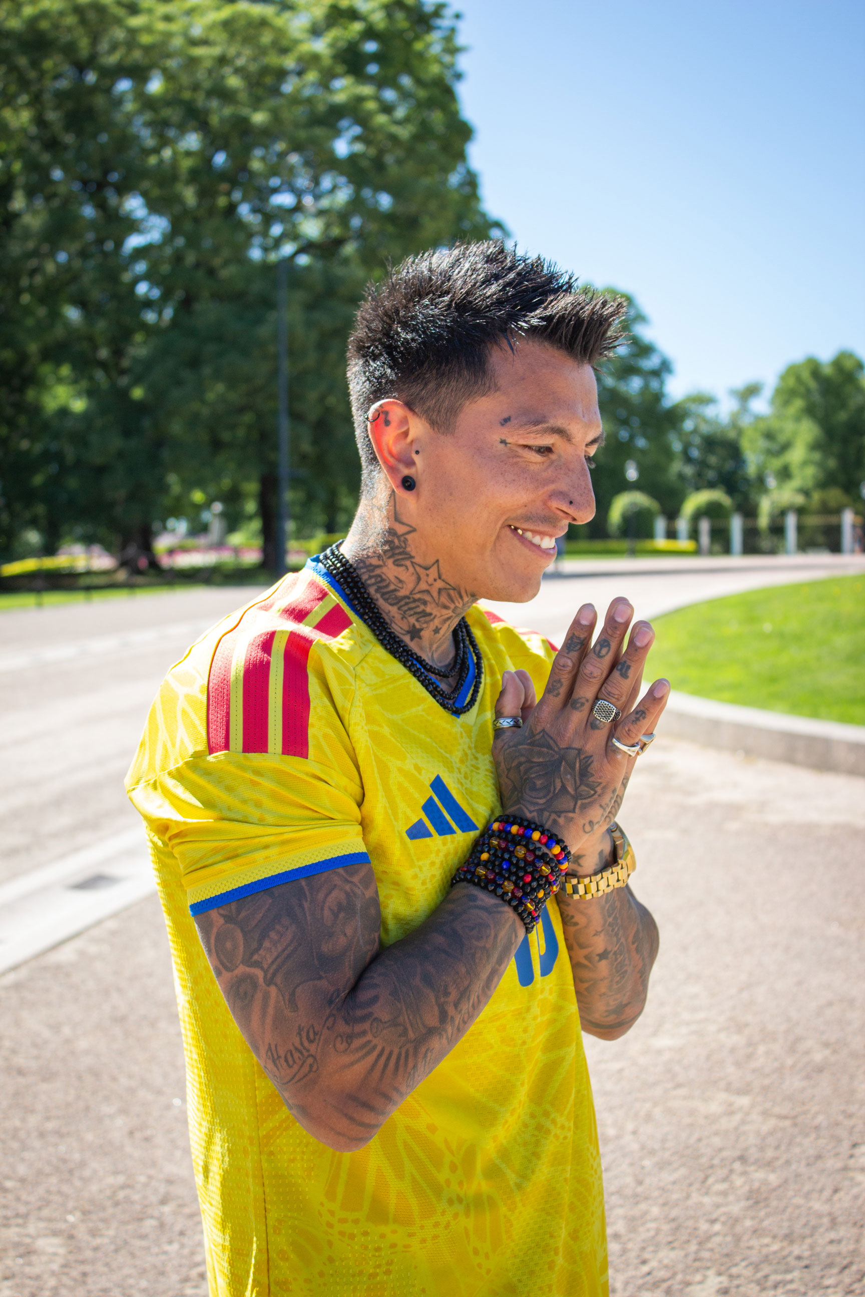Foto: Mann med tatoveringer, mørk piggsveis og gul fotballtrøye står med hendene mot hverandre, som i en bønn. Hodet er litt bøyt og han har en gulltann i smilet sitt.
