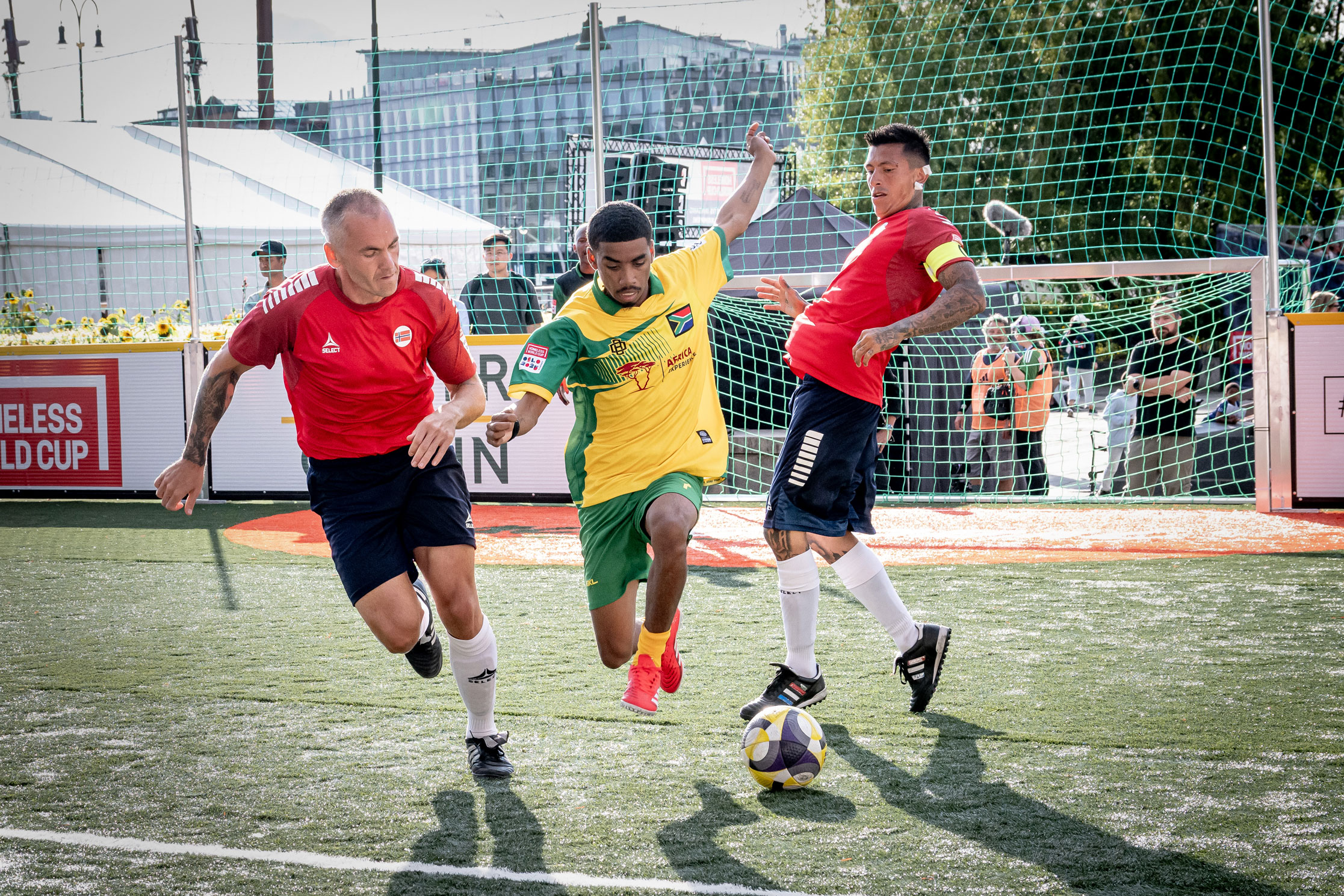 Foto: To spillere i rød drakt og en i midten i gul og grønn kjemper om en fotball på banen.