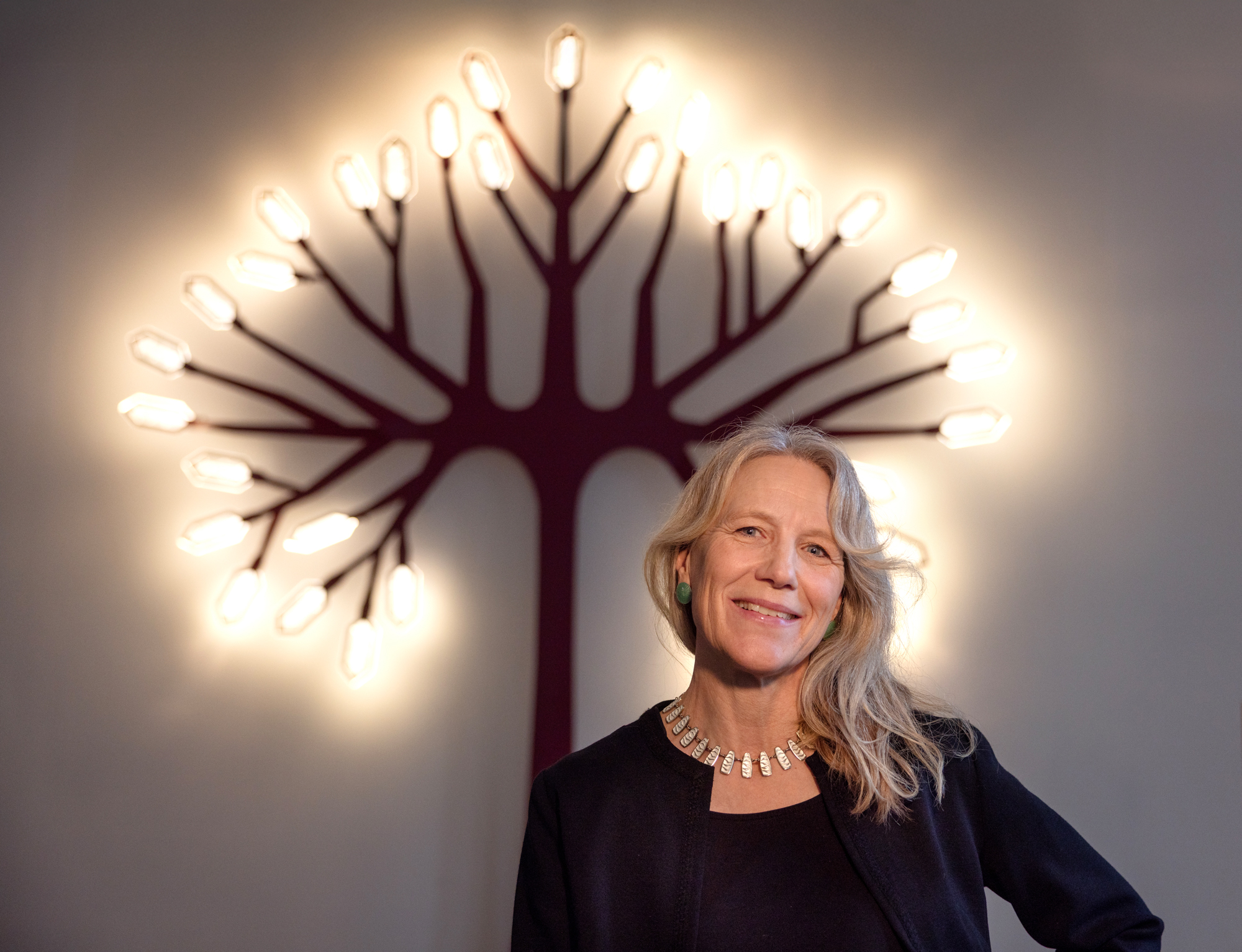 Foto: Kvinne i 50-årene med blondt hår står innendørs, foran et kunstverk som er et rødt tre i metall med mange grener. Ytters er det "blader" i form av sekskantede ledlys som skinner. Hun er festkledd i svart drakt med tilhørende jakke og har et stort, hvitt emaljesmykke rundt halsen.
