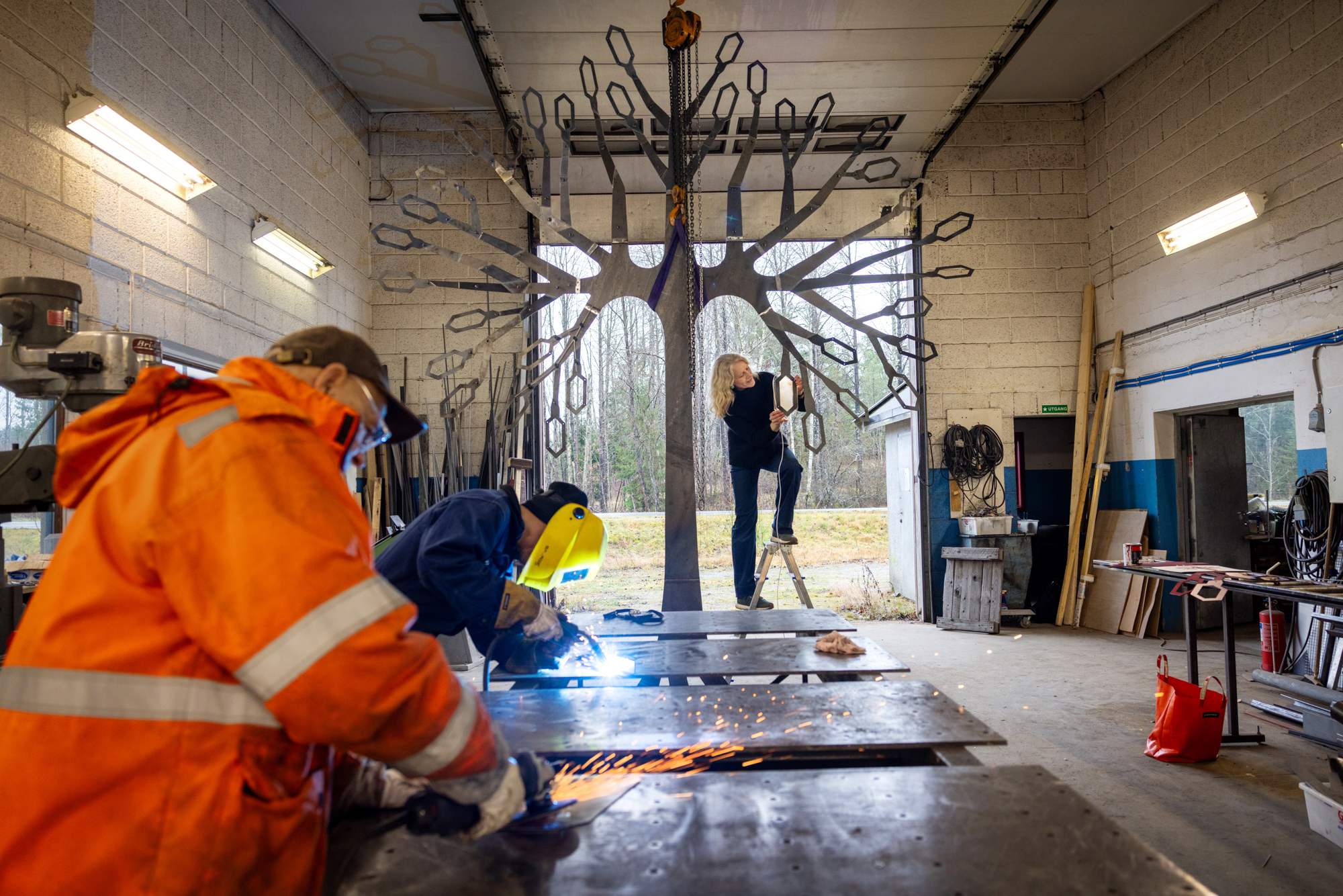 Foto: Fra en industrihall. En blond dame står på en gardintrapp ved et tre i metall. Ved et bord står to menn og sveiser.