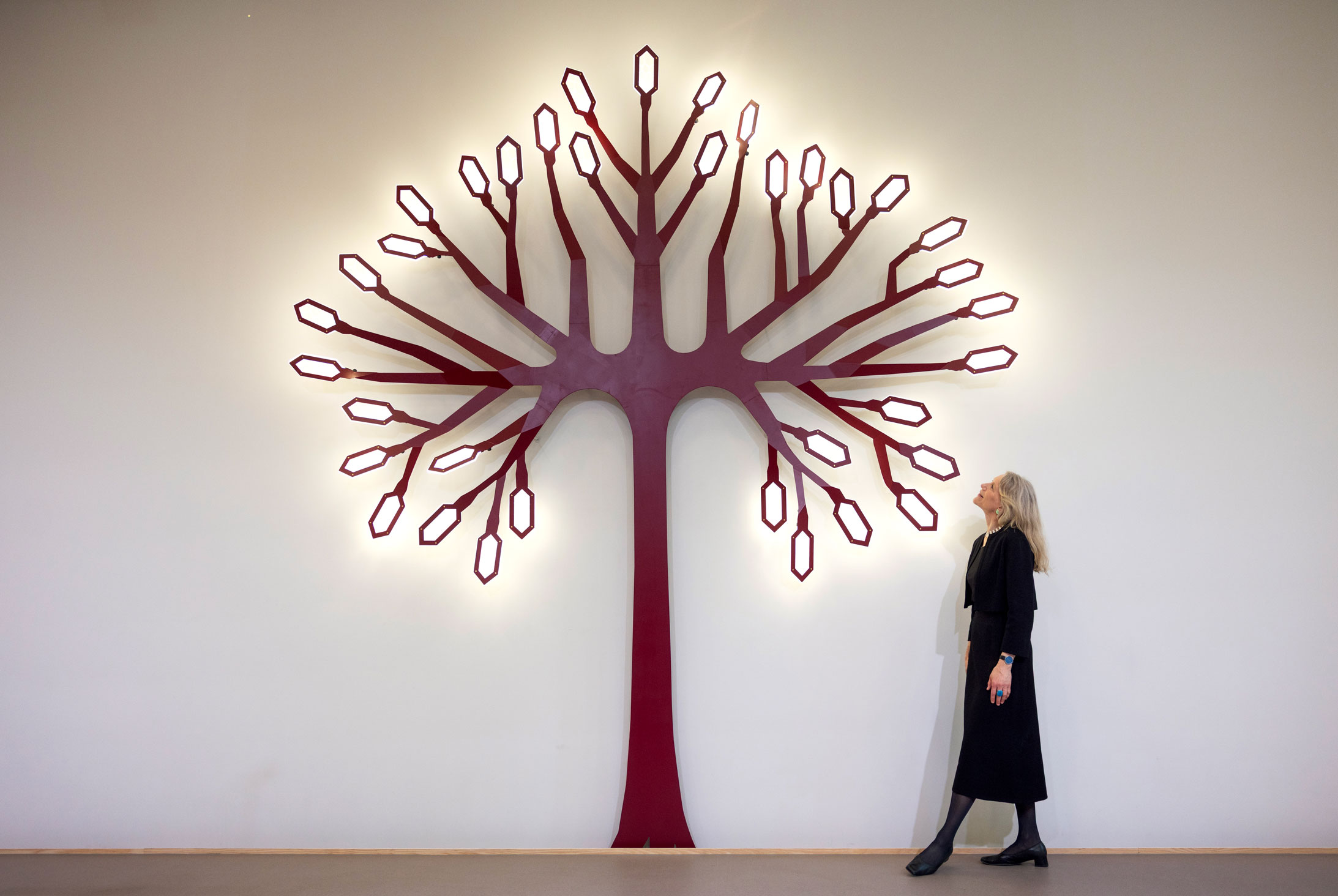 Foto: Kvinnen i svart drakt kikker opp på et stort, rødt tre i metall som er fastmontert i en hvit vegg. Det er tente lys på det som skal forestille blader.