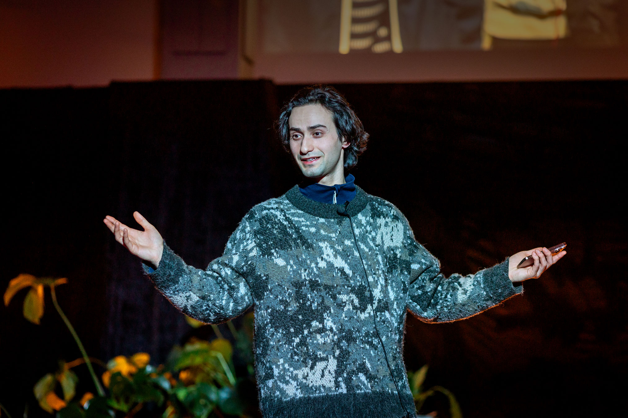 Foto: Mann med mørkt hår og brune øyne holder begge armene ut. Han står på en mørk scene og har på seg en ullgenser med mønster i ulike grønnfarger.