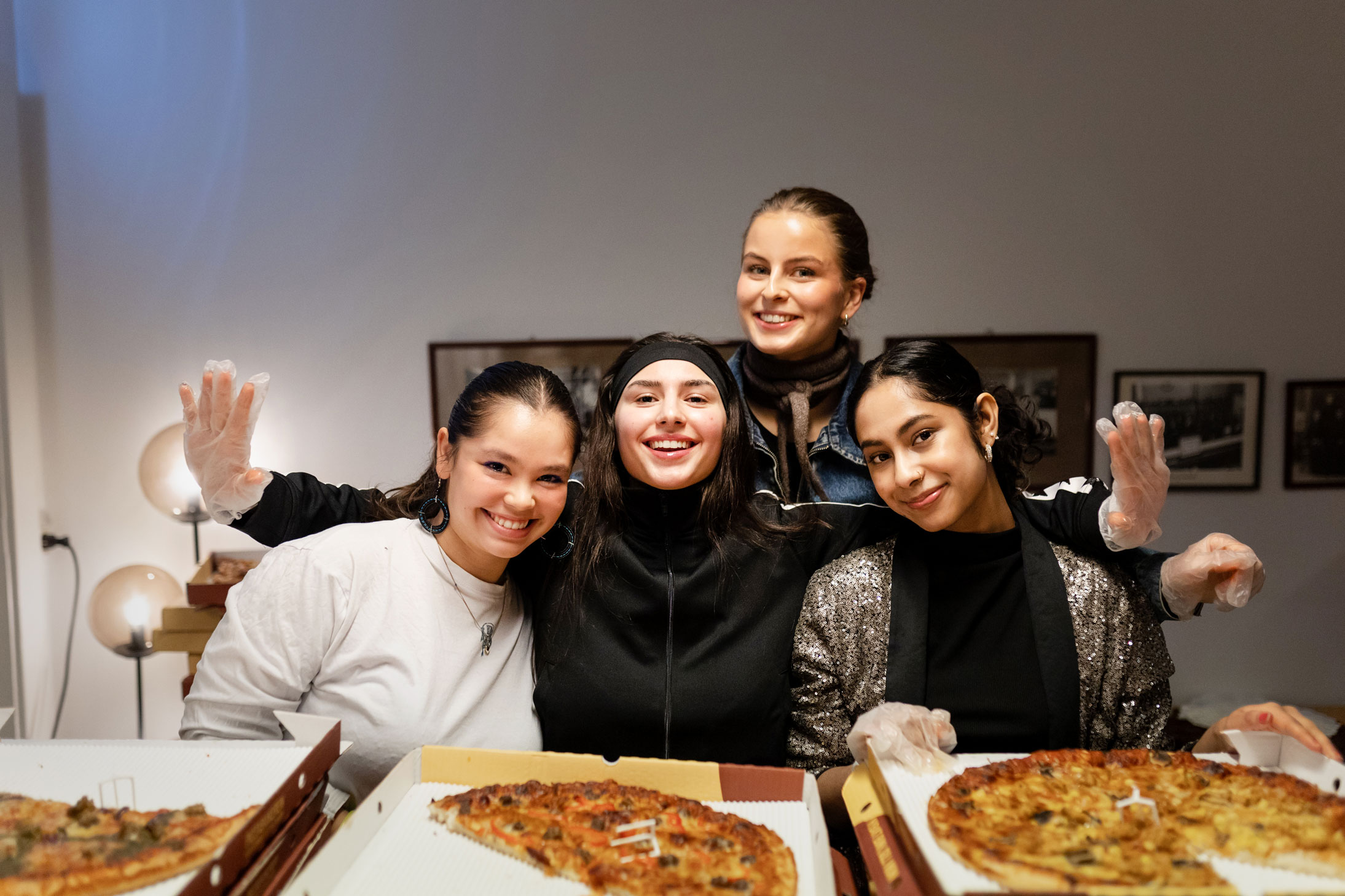 Foto: Fire smilende unge jenter står bake en disk full av esker med pizza.
