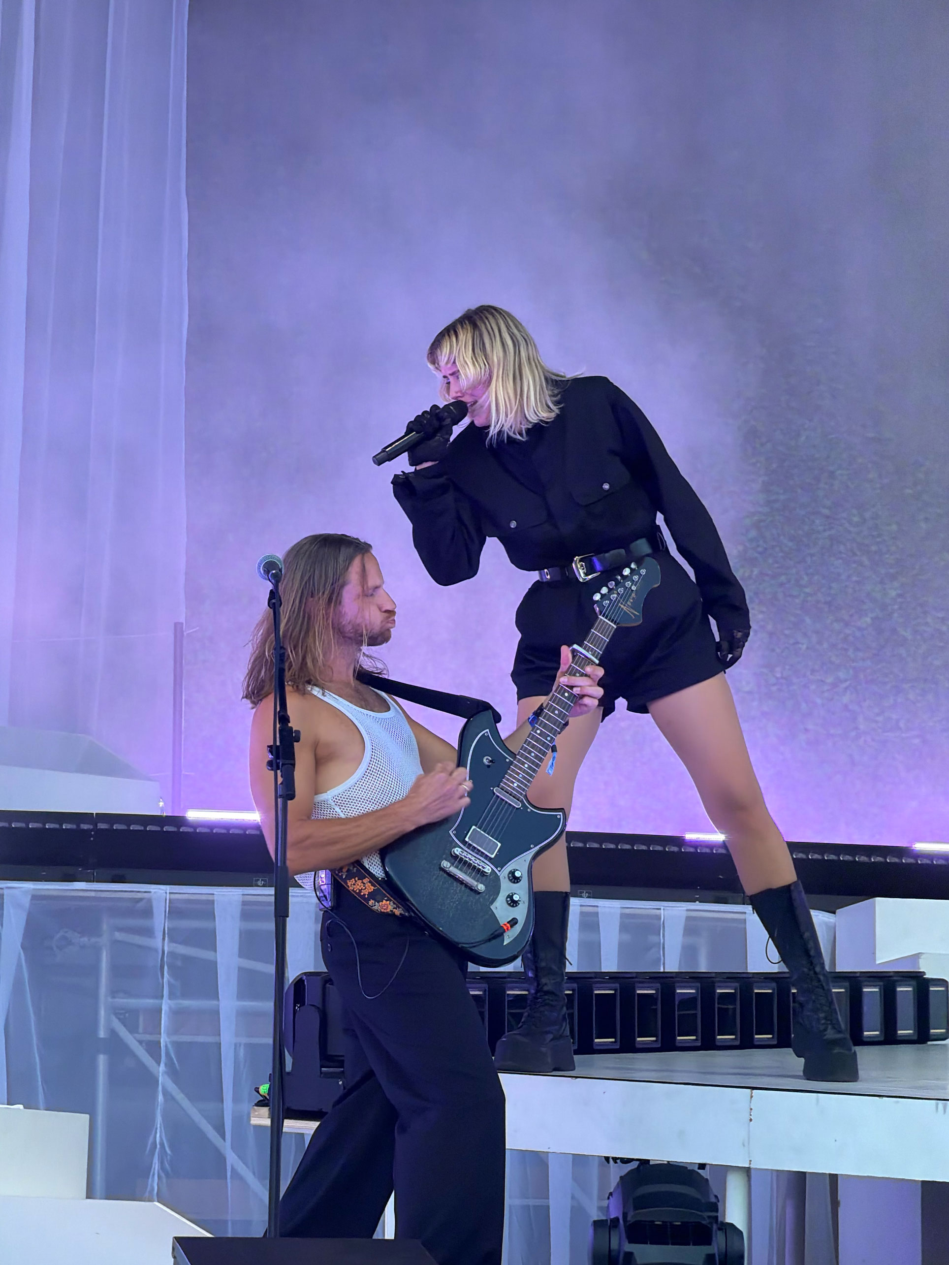 Foto: Blond kvinne i 30-årene står på en scene iført en svart, kort kjeledress med bare legger og svarte boots. Hun synger i en mikrofon vendt mot en mannlig gitarist.
