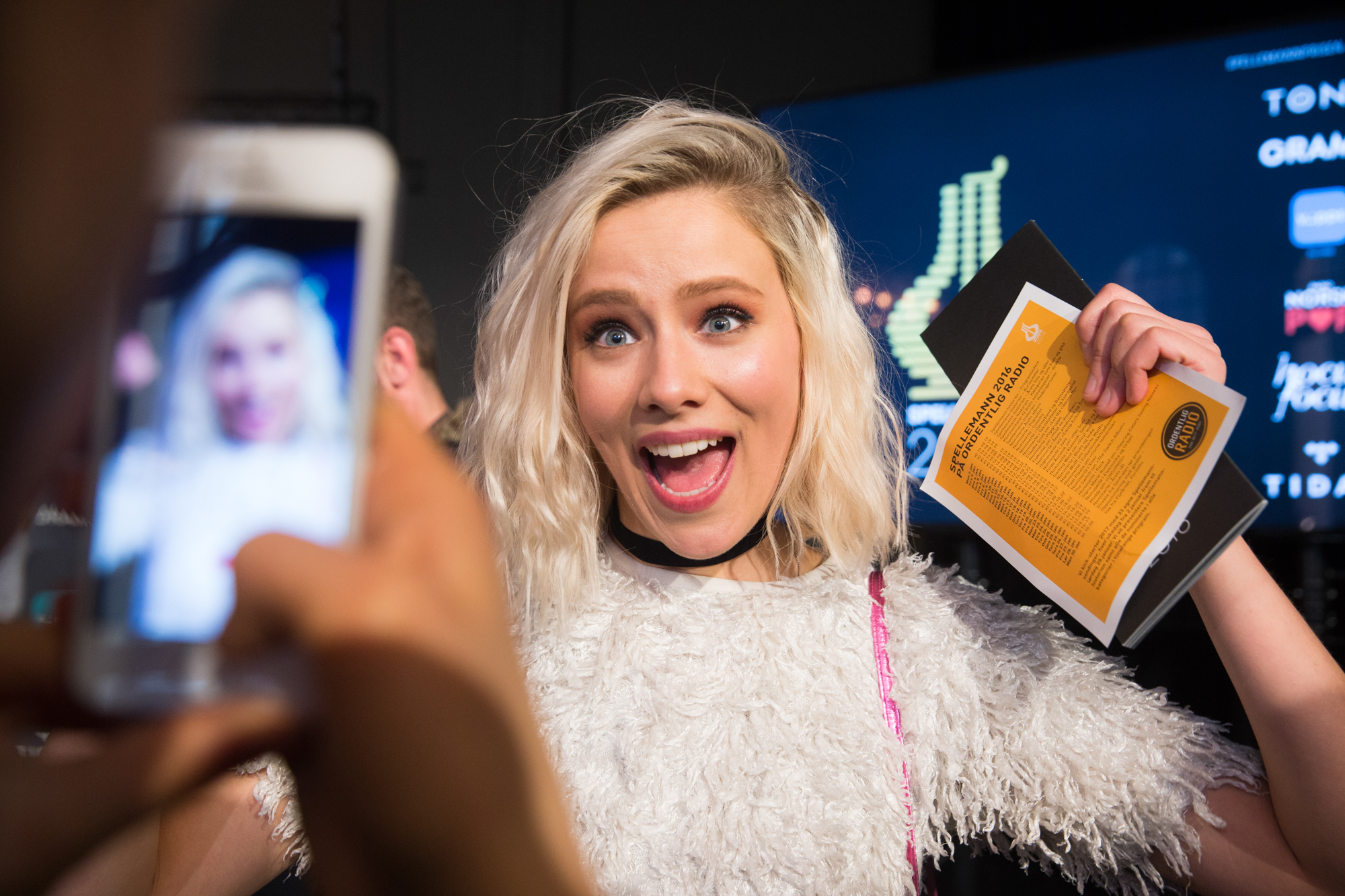 Foto: Ung, blond jente med blå øyne smiler til kamera idet noen tar bilde av henne gjennom en Iphone. Hun har på seg en hvit, hårete topp og på veggen bak er det logoen til Spellemann.
