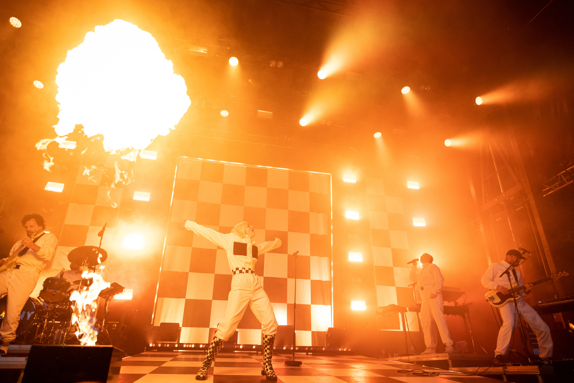 Foto: Et band på scenen med pyro effekter og sjakkrutete teppe i svart-hvitt i bakgrunnen. En kvinne i front strekker ut armene bakover.