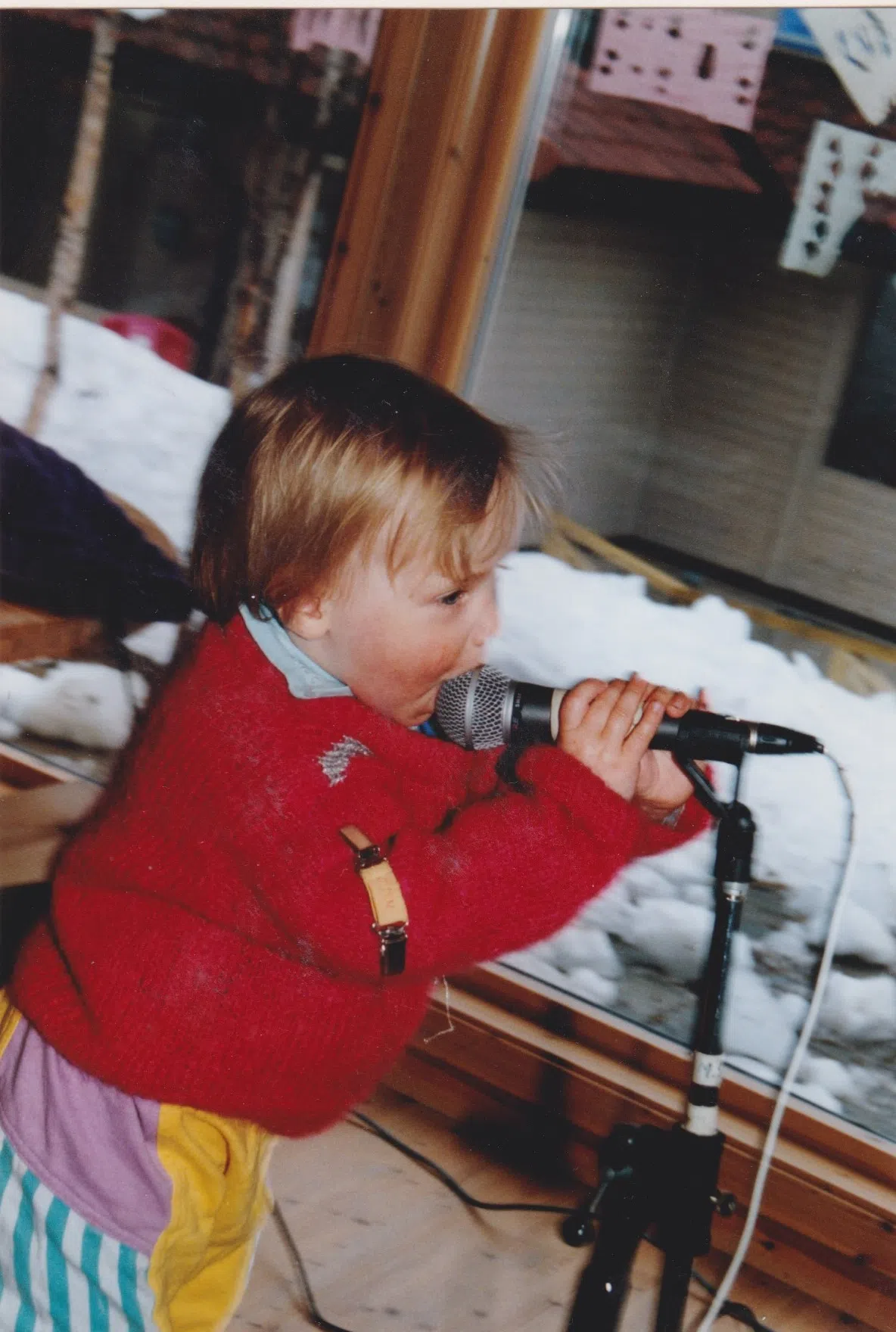 Foto: 2-åring i rød strikkehenser og fargerike bukser synger i en mikrofon. Det er litt snø på terrassen utenfor.