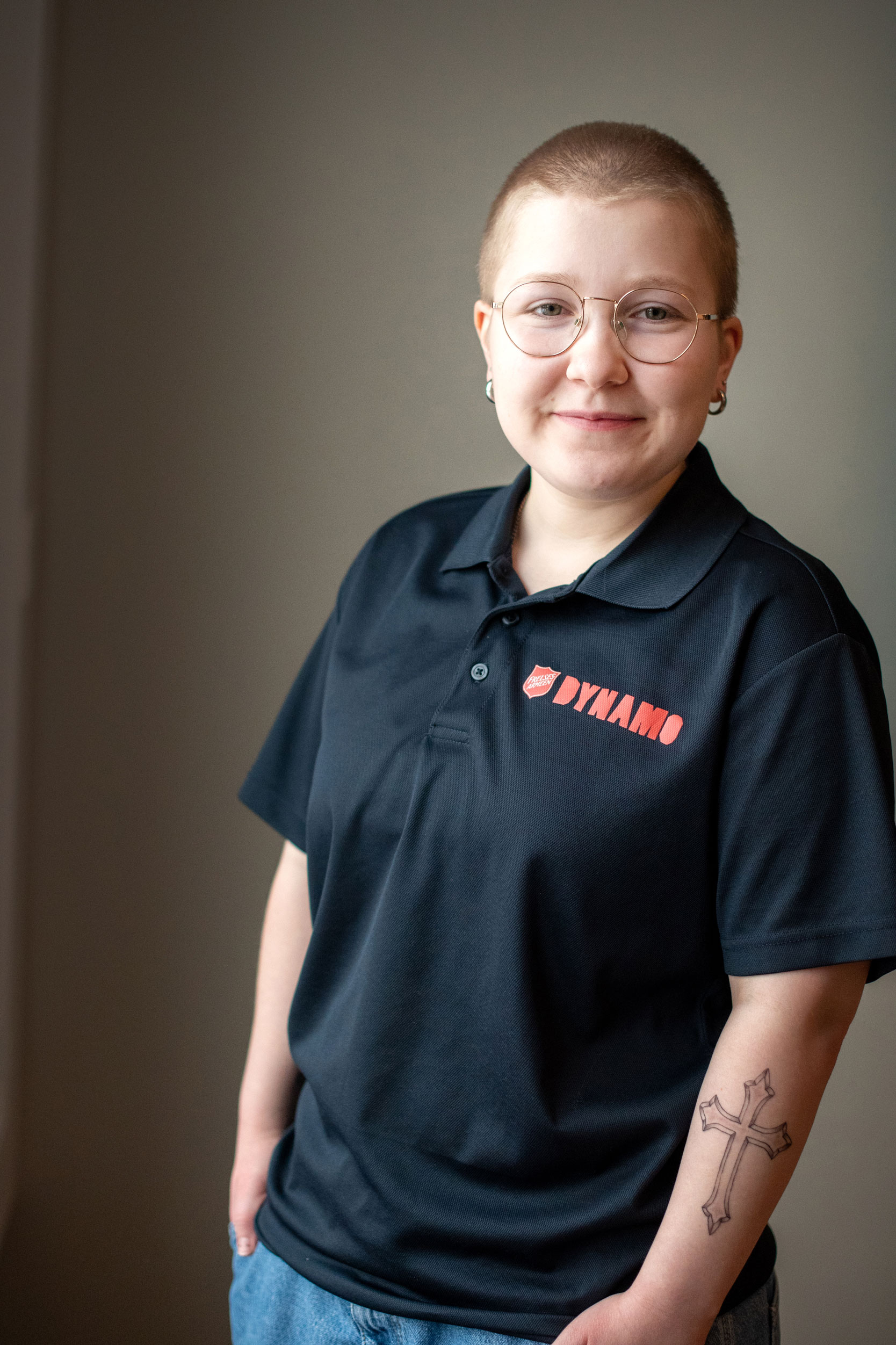 Foto: Kvinne med nesten helt glattbarbert, blondt hår lener seg inntil en vegg. Hun har runde briller med gullkant, og har på seg en mørkeblå piquetrøye der det står Frelsesarmeens logo og DYNAMO med store bokstaver. På venstre arm har hun tatovert inn et stort kors.