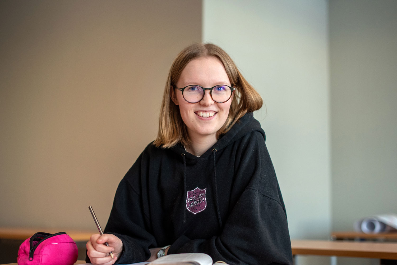 Foto: Kvinne med halvlangt, mellomblondt hår og briller med mørk kant smiler til kamera. Hun sitter i et klasserom og holder en blyant. Ved siden av ligger et rosa penal og hun har en bok oppslått på pulten.
