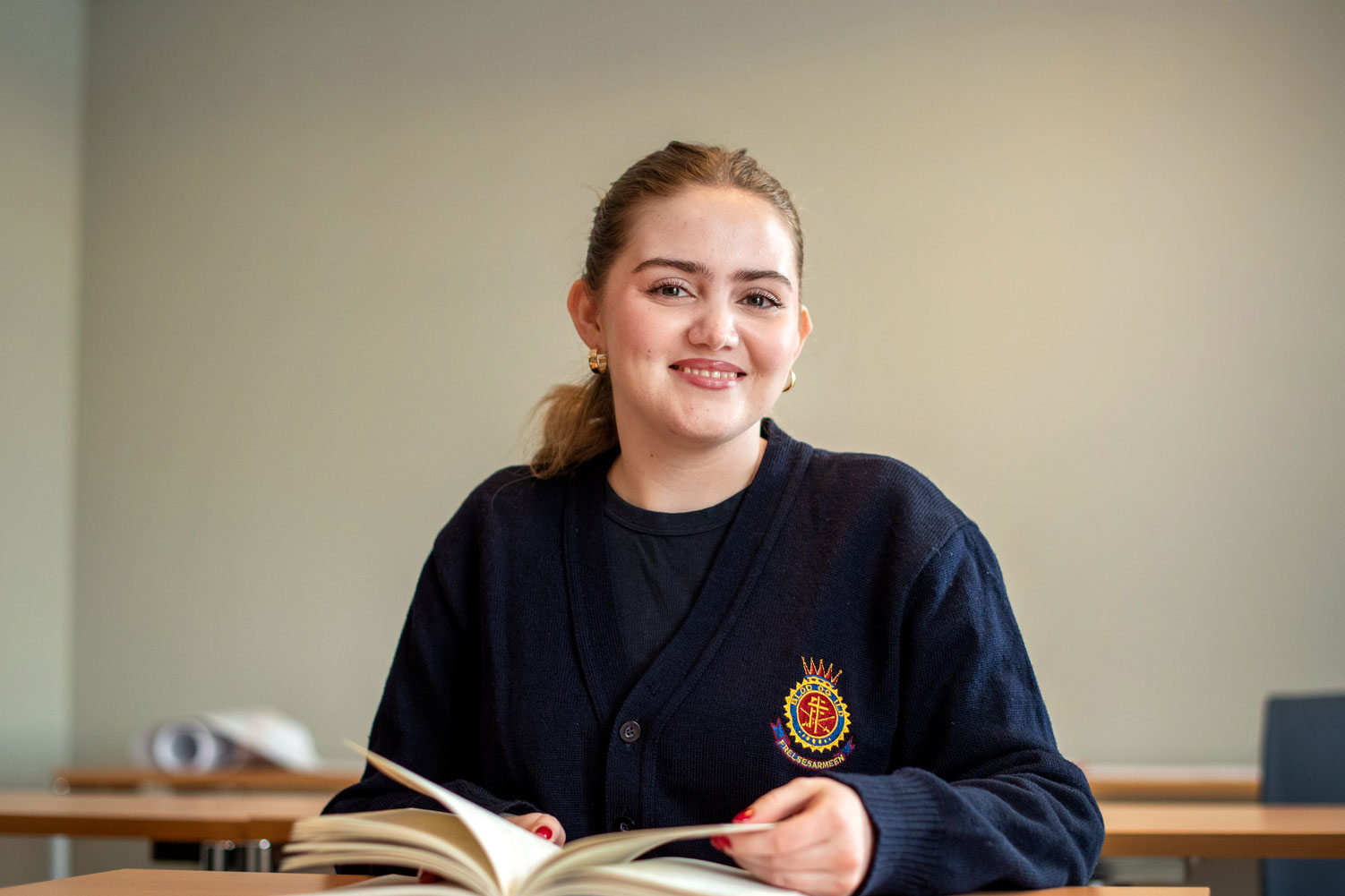 Foto: Kvinne tidlig i 20-årene med brune øyne og brunt hår i hestehale sitter ved en pult i et klasserom. Hun holder en oppslått bok i hendene.