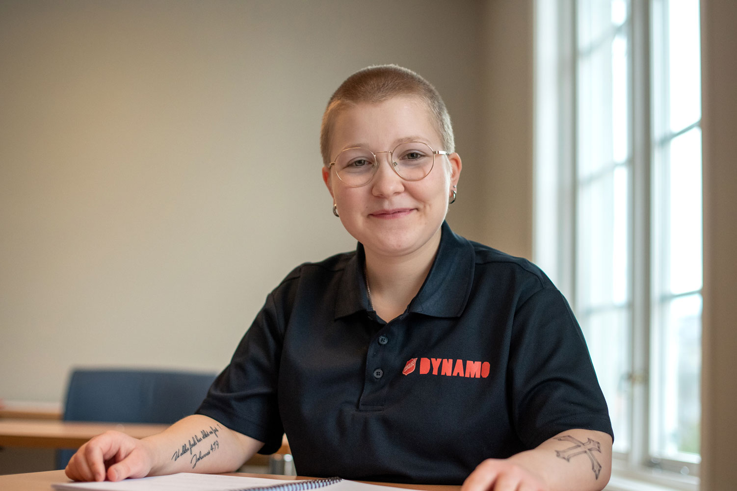 Foto: Kvinne med nesten helt glattbarbert, blondtsitter ved en pult i et klasserom. Hun har runde briller med gullkant, og har på seg en mørkeblå piquetrøye der det står Frelsesarmeens logo og DYNAMO med store bokstaver. Hun har tatoveringer på begge underarmene.