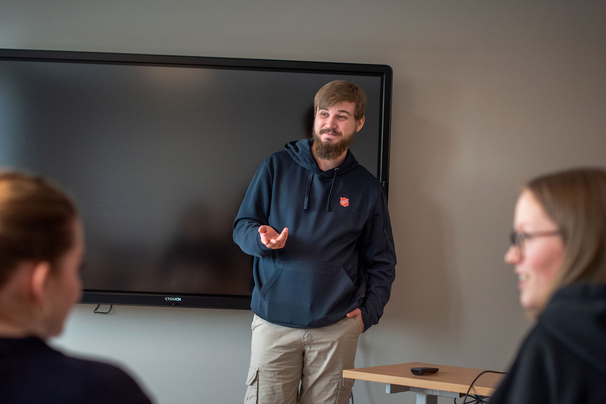 Foto: Mann i midten av 20-årene med skjegg og blå hettegenser står foran i et klassserom med TV-skjerm og underviser. På et par av stolene sitter det unge kvinner.
