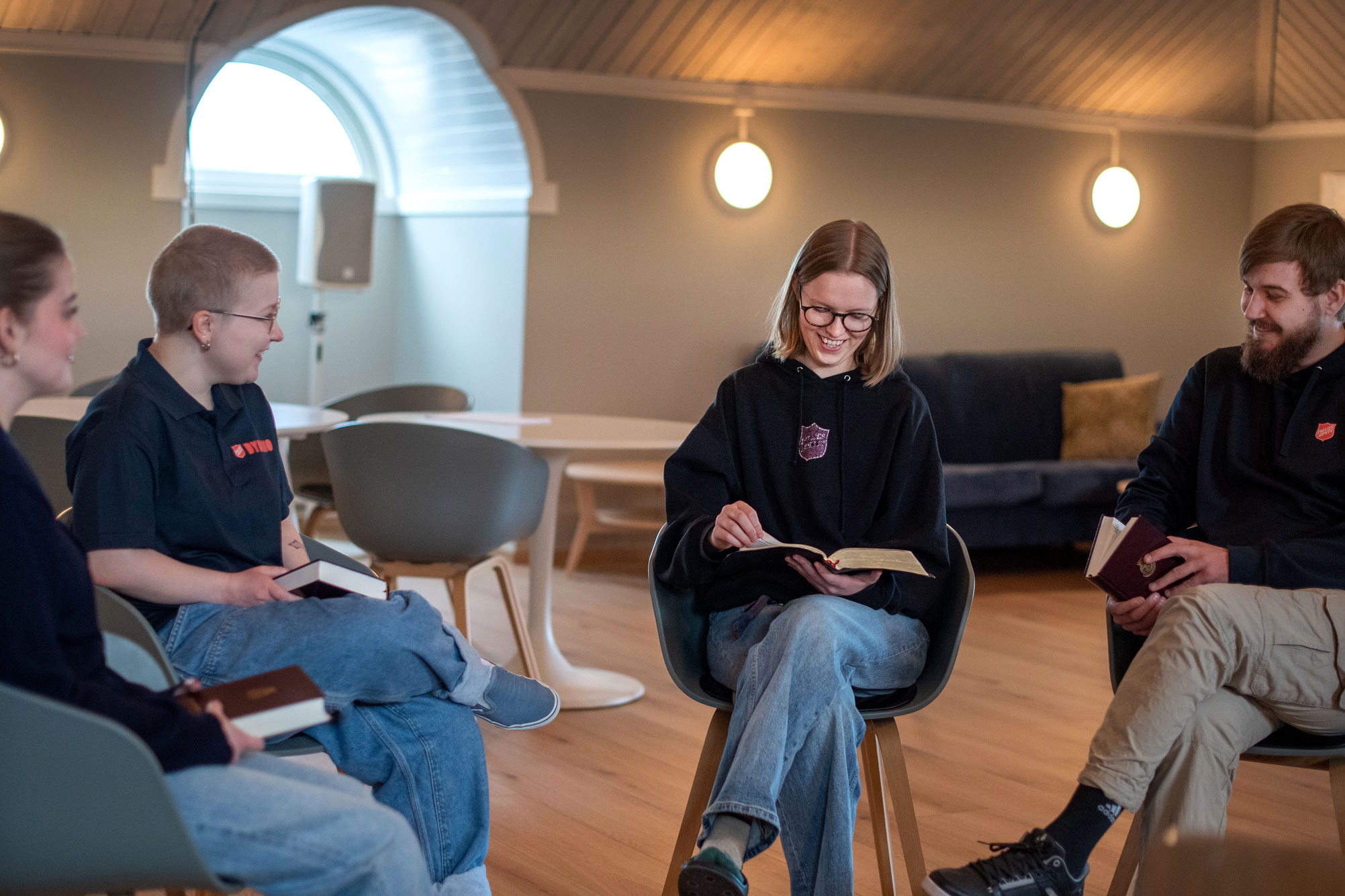 Foto: Mann og tre kvinner sitter på stoler i en sirkel. De har hver sin bok i hendene. Rommet har litt skråtak og i den ene enden er det et stort, buet vindu med hvite karmer. Det åpne rommet er ellers grått på veggene og møblert med sofa, bord og stoler..