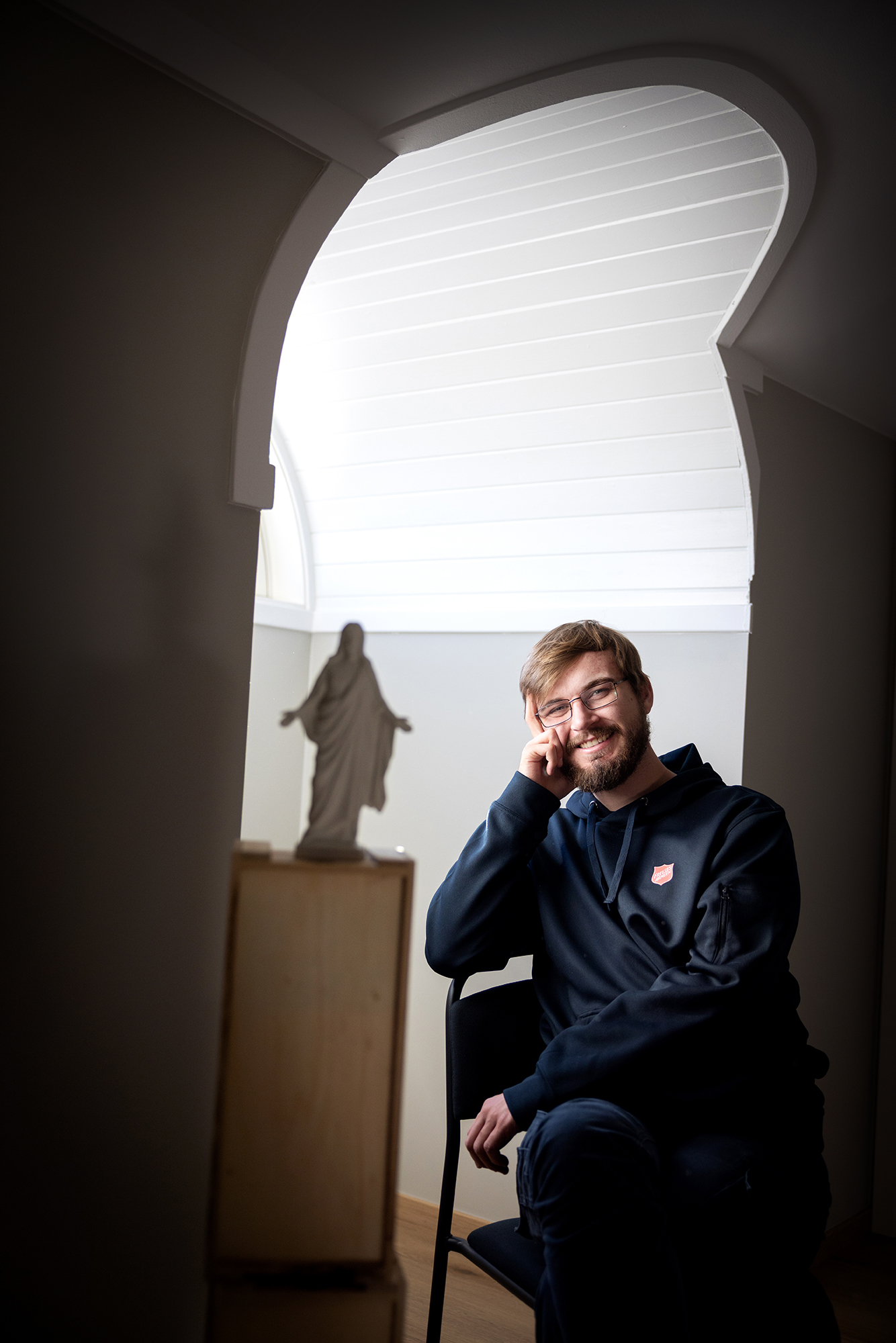 Foto: Mann i midten av 20-årene med briller og skjegg, sitter på en stol i et vindu der det står en Kristus-figur på et bord ved siden av. Han har på seg blå hettegenser med logo fra Frelsesarmeen. Han støtter hodet sitt i høyre hånd.
