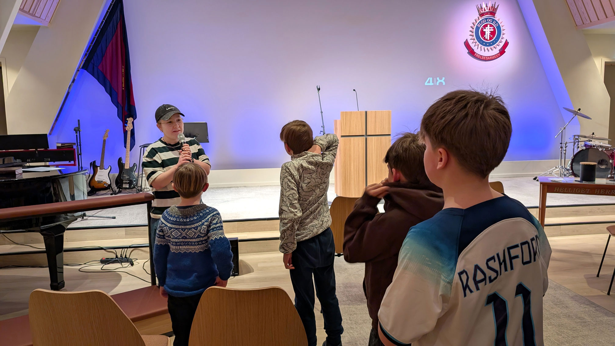 Foto: Ung kvinne med caps og stripene genser står inne i et kirkerom med en gruppe på fire gutter.