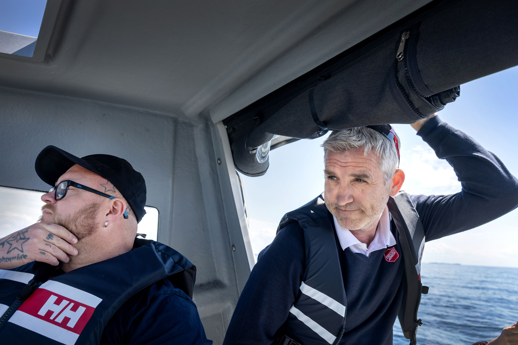Foto: To menn inne i båten med livvester på. Han med caps, briller og tatoveringer styrer. Han andre med grått hår og skjold fra Frelsesarmeen på genseren holder seg fast i den opprullede båtkalesjen.