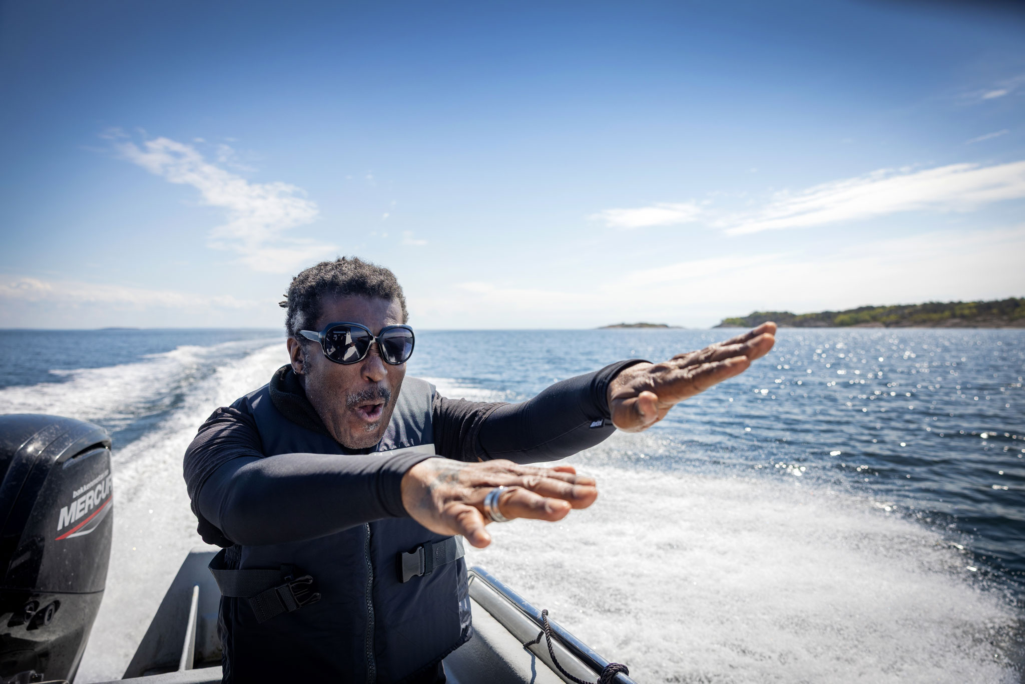 Foto: Svart mann med krøller og mørke solbriller står bak på en båt. Han holder hendene fram, som om han surfer.