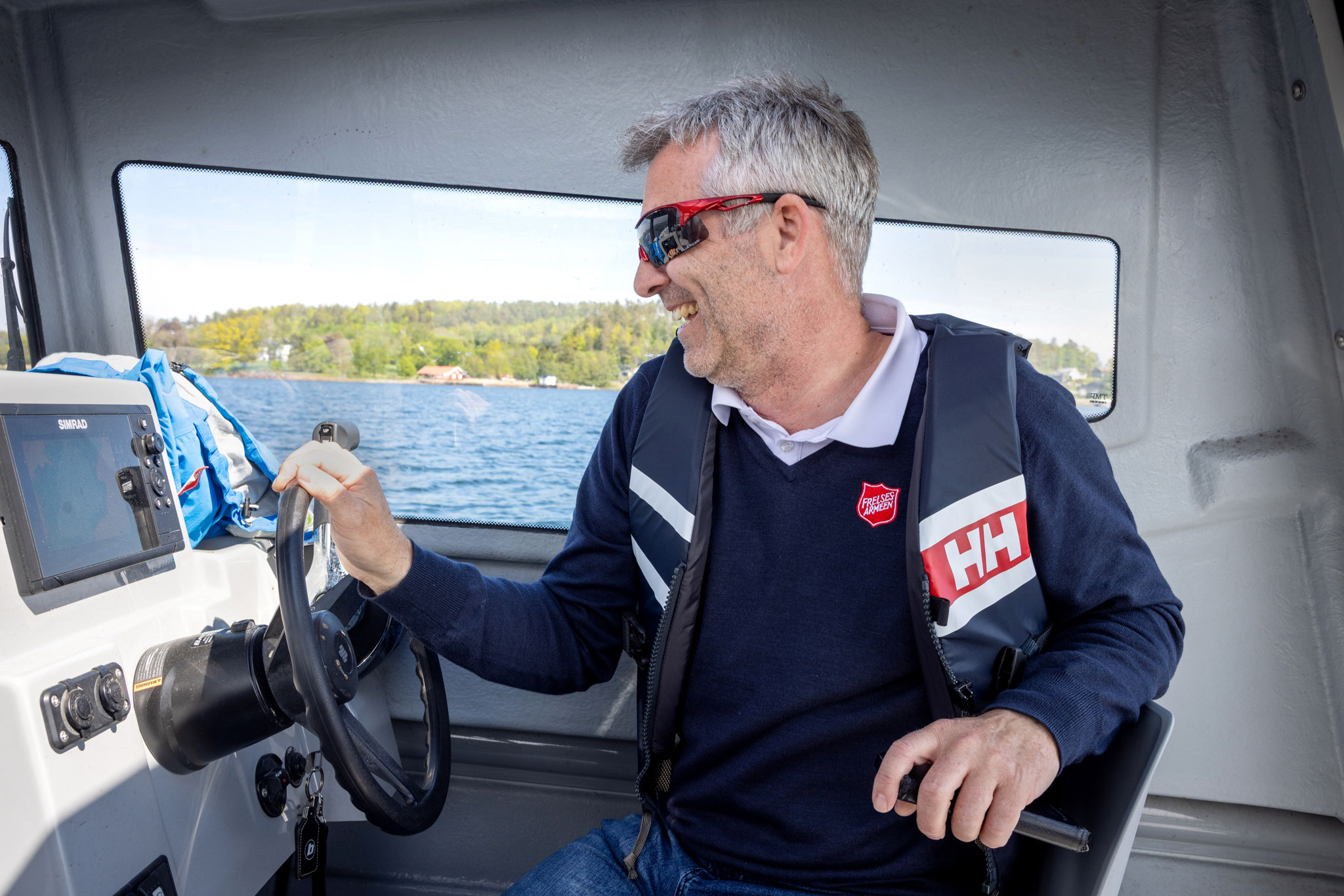 Foto: Mann med Frelsesarme-skjold på genseren styrer båten. Han har på raske. røde briller og blå, hvit og rød redningsvest fra HH.