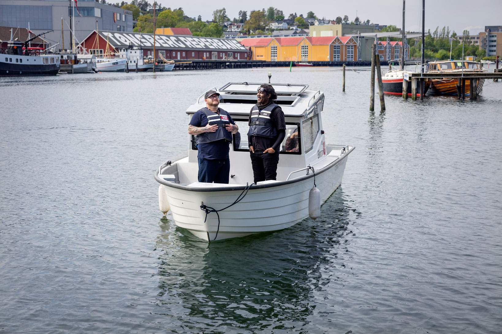 Foto: To menn med redningsvester står foran i en liten, hvit plastbåt på sjøen inne i et havneområde.