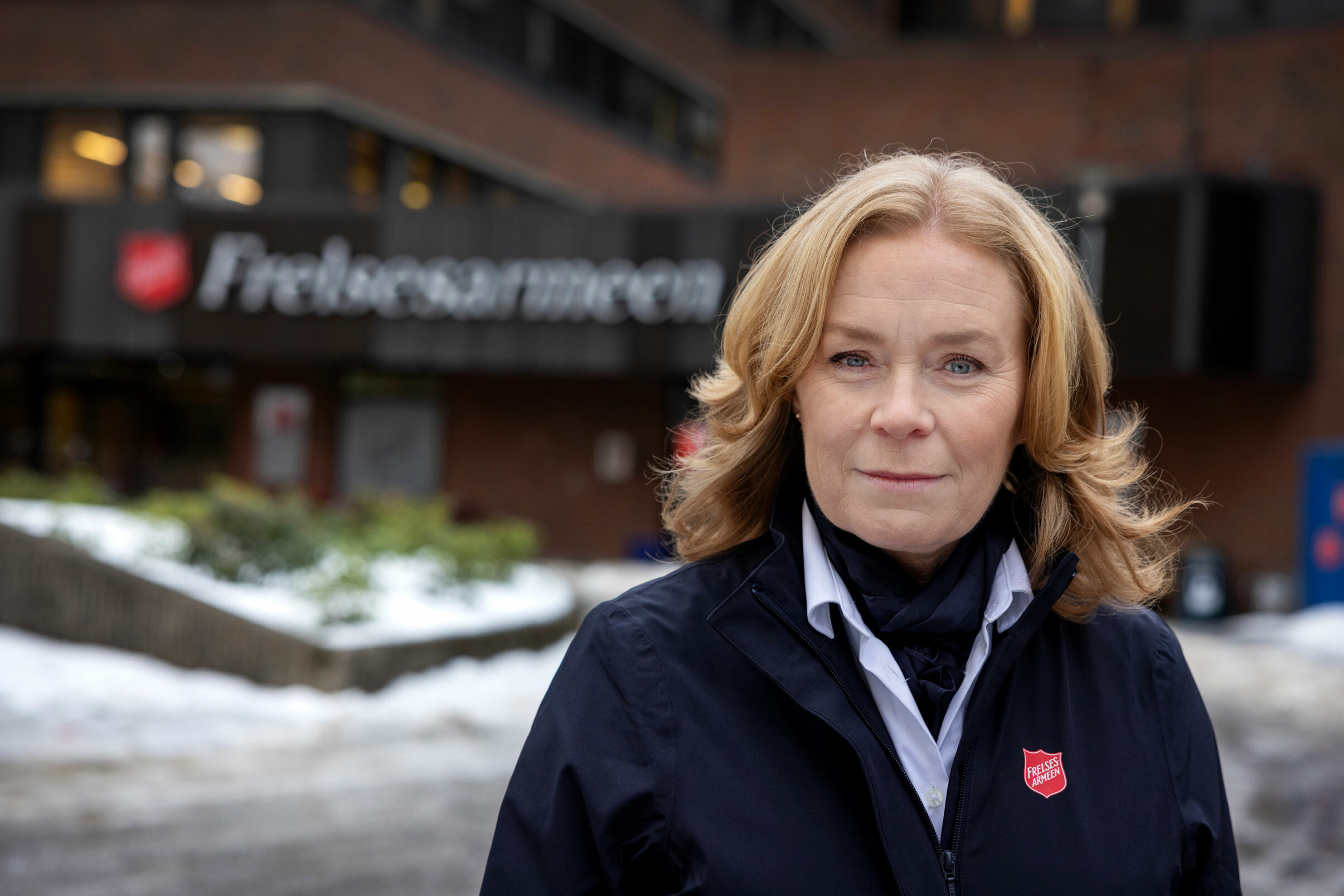 Kvinne i uniform står utenfor et stort bygg med Frelsesarmeens logo utenpå. Litt snø på bakken.