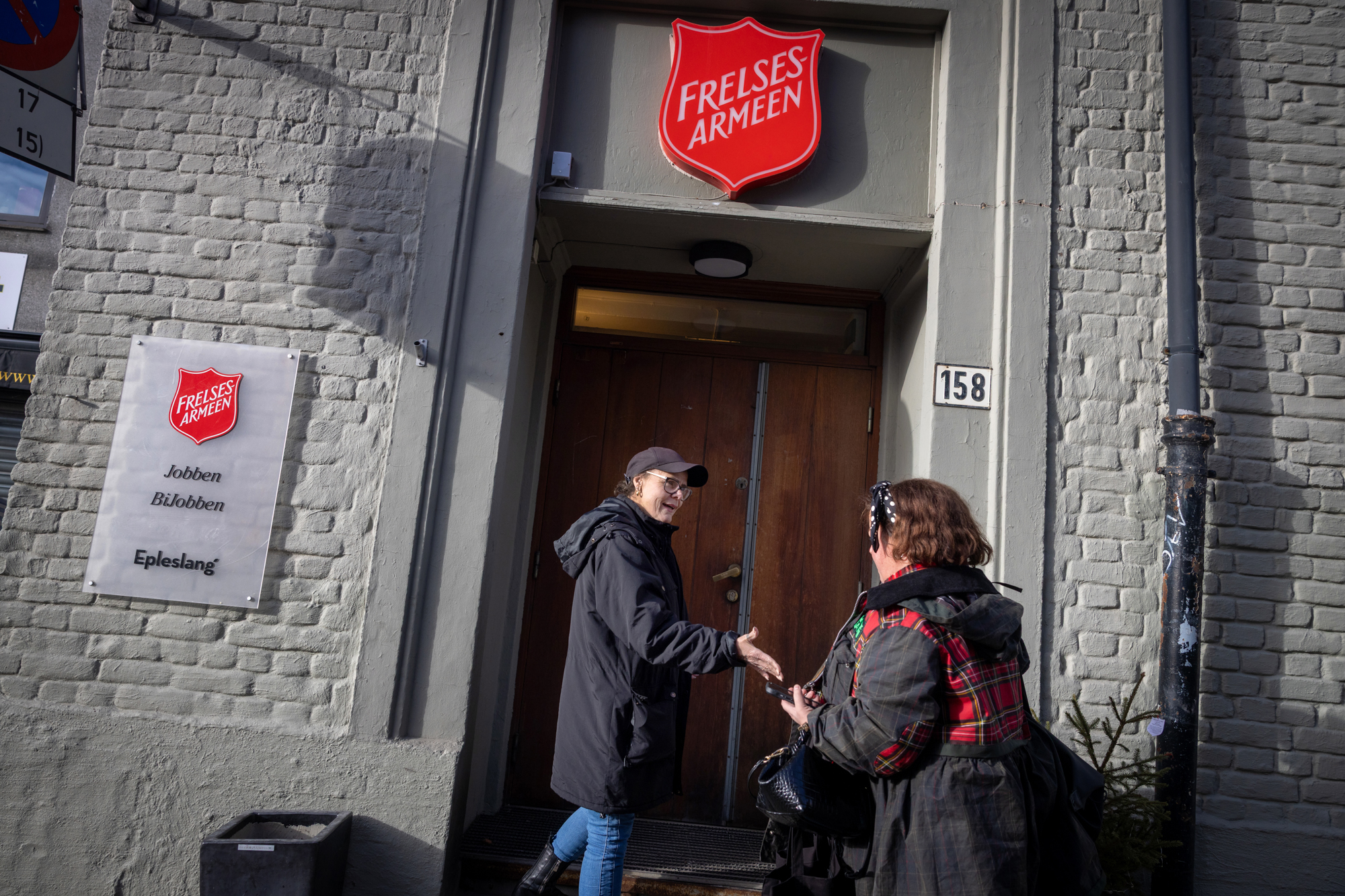 Foto: To kvinner håndhilser på hverandre foran en dør inn til et murbygg med skilt med logo fra Frelsesarmeen skrudd inn i fasaden.