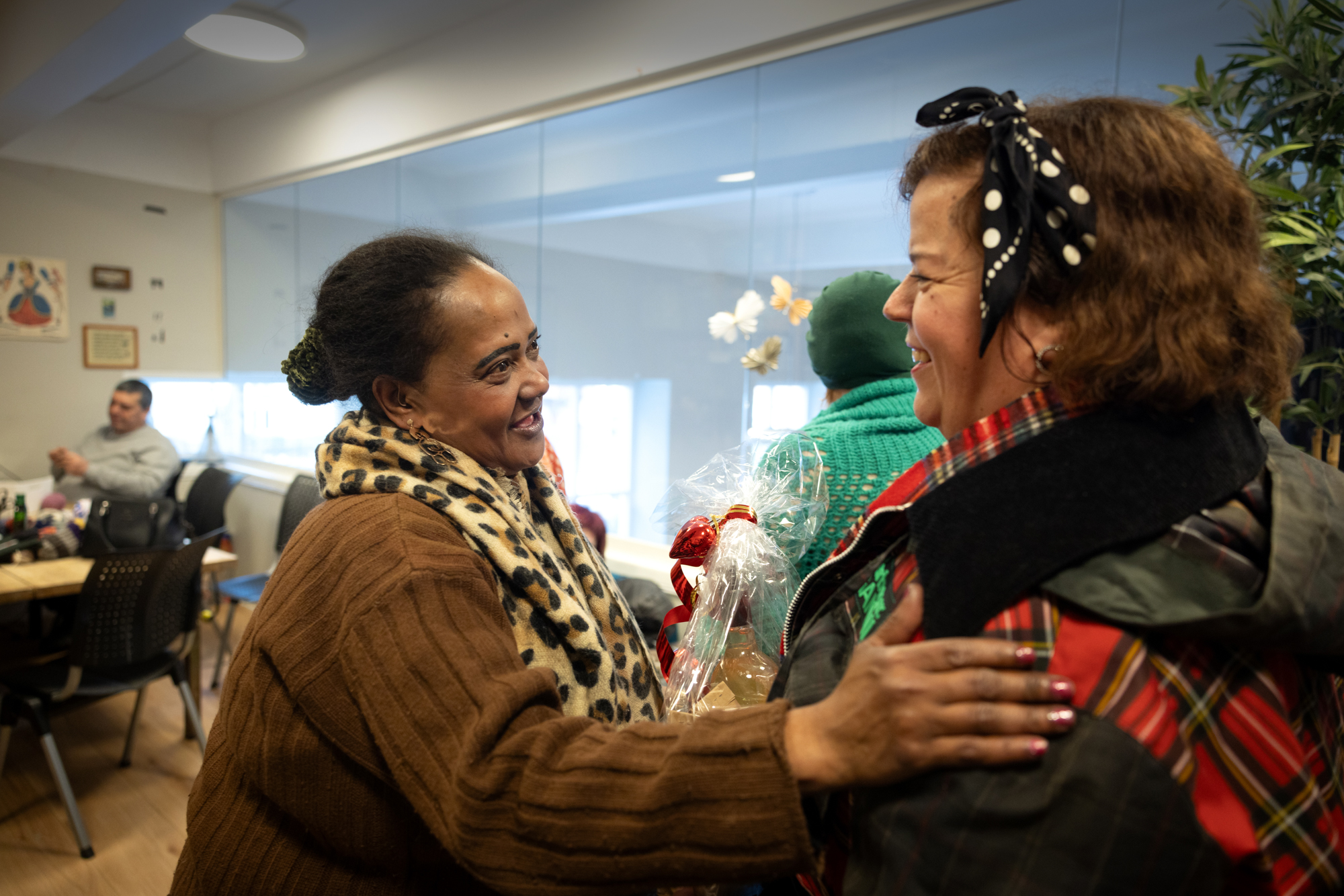 Foto: Svart kvinne i brun genser og et leopardmønstrete skjerf i halsen, holder armen på skulderen til kvinne med mørkt hår og grønn jakke med rødrutete detaljer. Begge smiler.