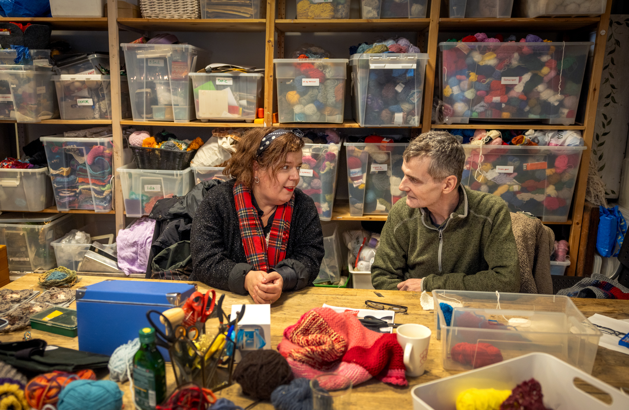 Foto: Inne på et hobbyrom med en masse gjennomsikte plastkasser med garn og andre materialer sitter en mann og en kvinne ved et bort. De ser på hverandre mens de snakker sammen.