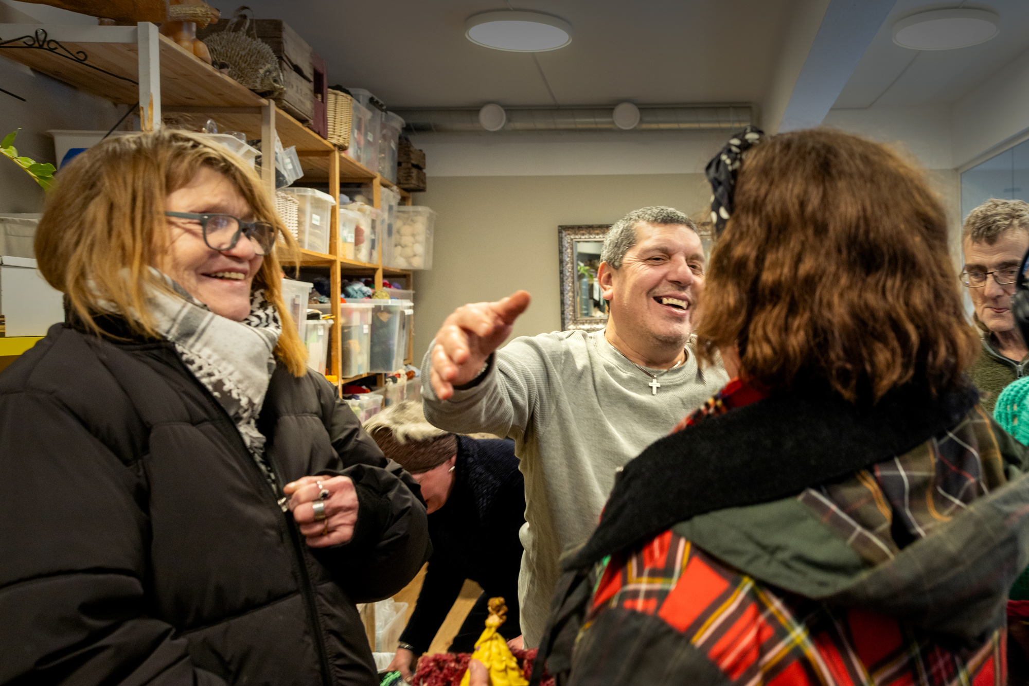 Foto: Kvinne med briller som smiler og iakttar en mann med grått hår og grå genser som strekker opp og ut for å gi damen i grønn jakke med rødrutete detaljer en klem.