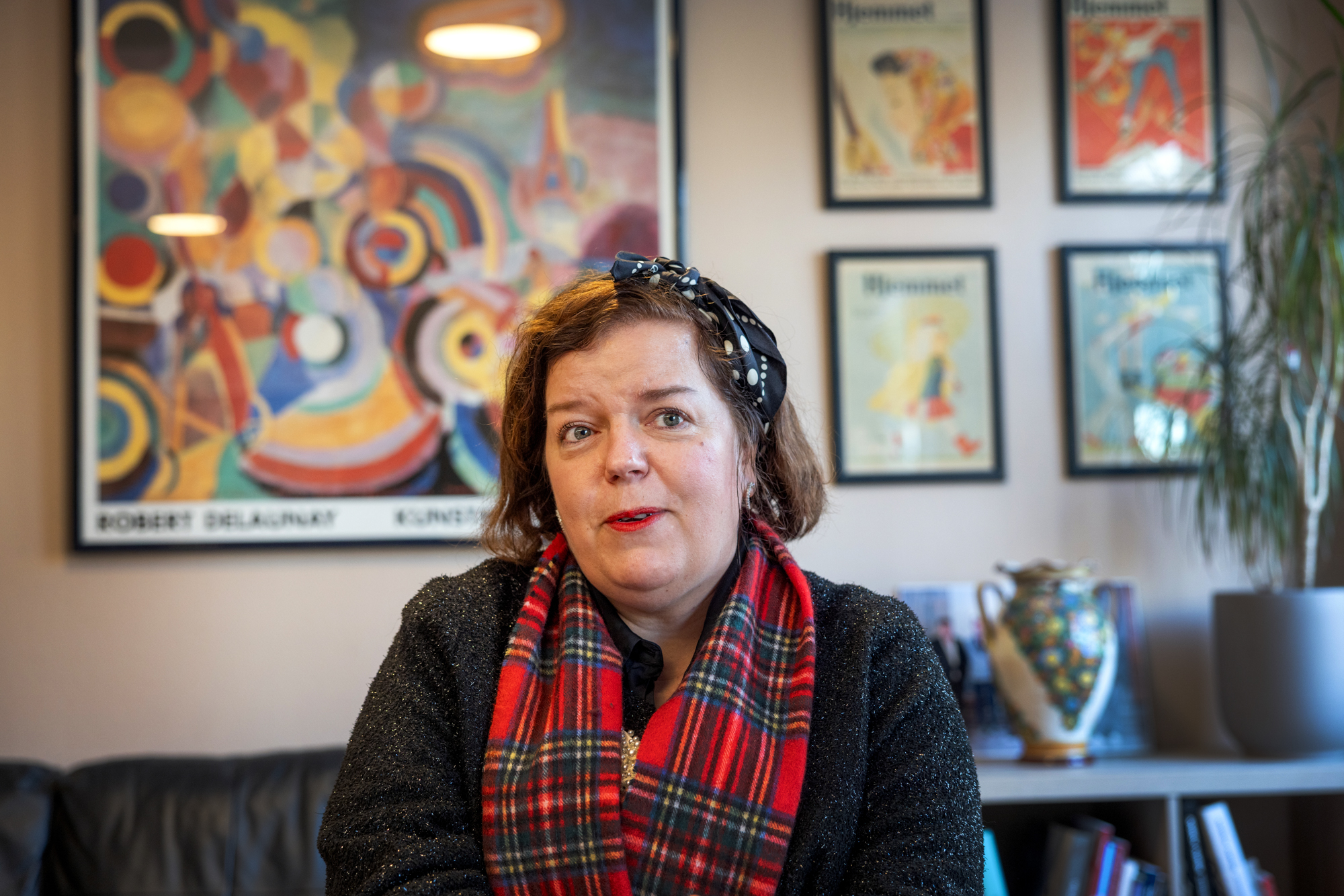 Foto: Kvinne med mørkt hår og rødrutete skjerf i halsen sitter i en sofa foran en vegg med kunstplakater.