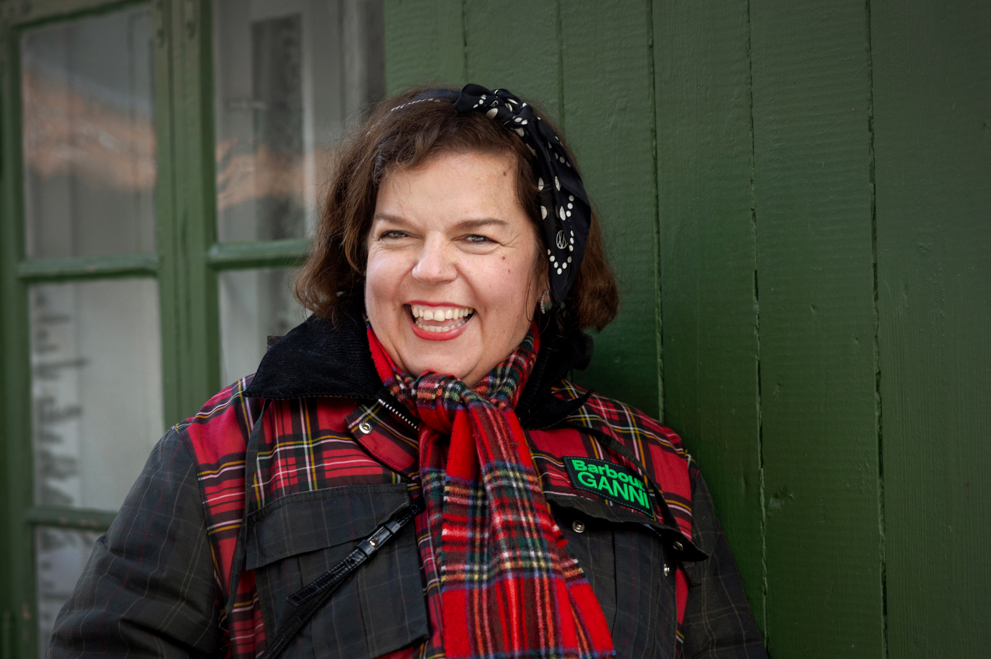 Foto: Kvinne midt i 40-årene med mørkt hår smiler foran en grønnmalt vegg. Hun har på seg en mørkegrønn jakke med rødrutete tøyfelt oppe over brystet og på lommene. Hun har et maken skjerf i halsen.