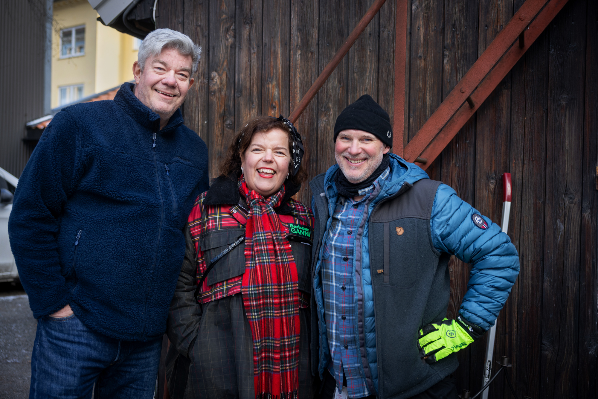Foto: En høy mann med grått hår står sammen med en dame og mann som er mye lavere og på samme høyde. De står foran en port i tre og mennene har på seg arbeidsklær.