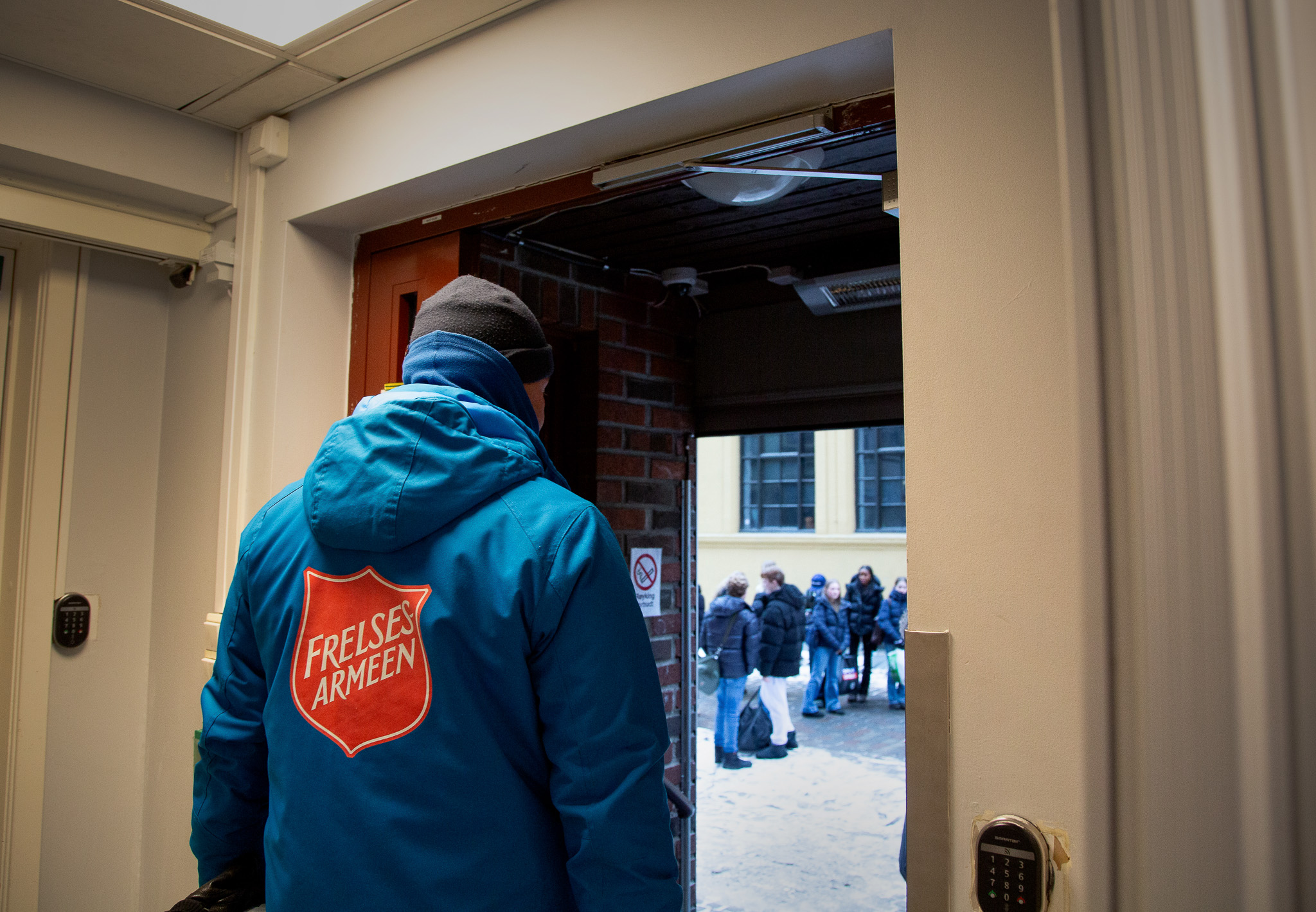 Mann med blå jakke med stor rød logo på står i inngangen til et bygg og ser ut. I bakgrunnen står flere skoleelever med poser i hendene.