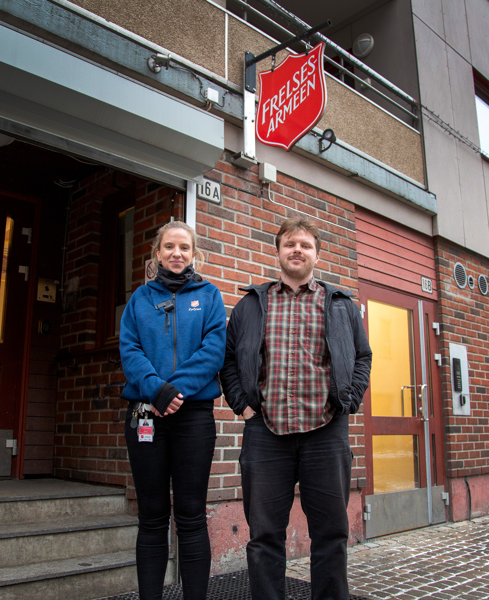 Foto: Blind kvinne i blå flisgenser med logo fra Frelsesarmeen står sammen med en mann i rutete skjorte utenfor en rød mursteinsbygning.