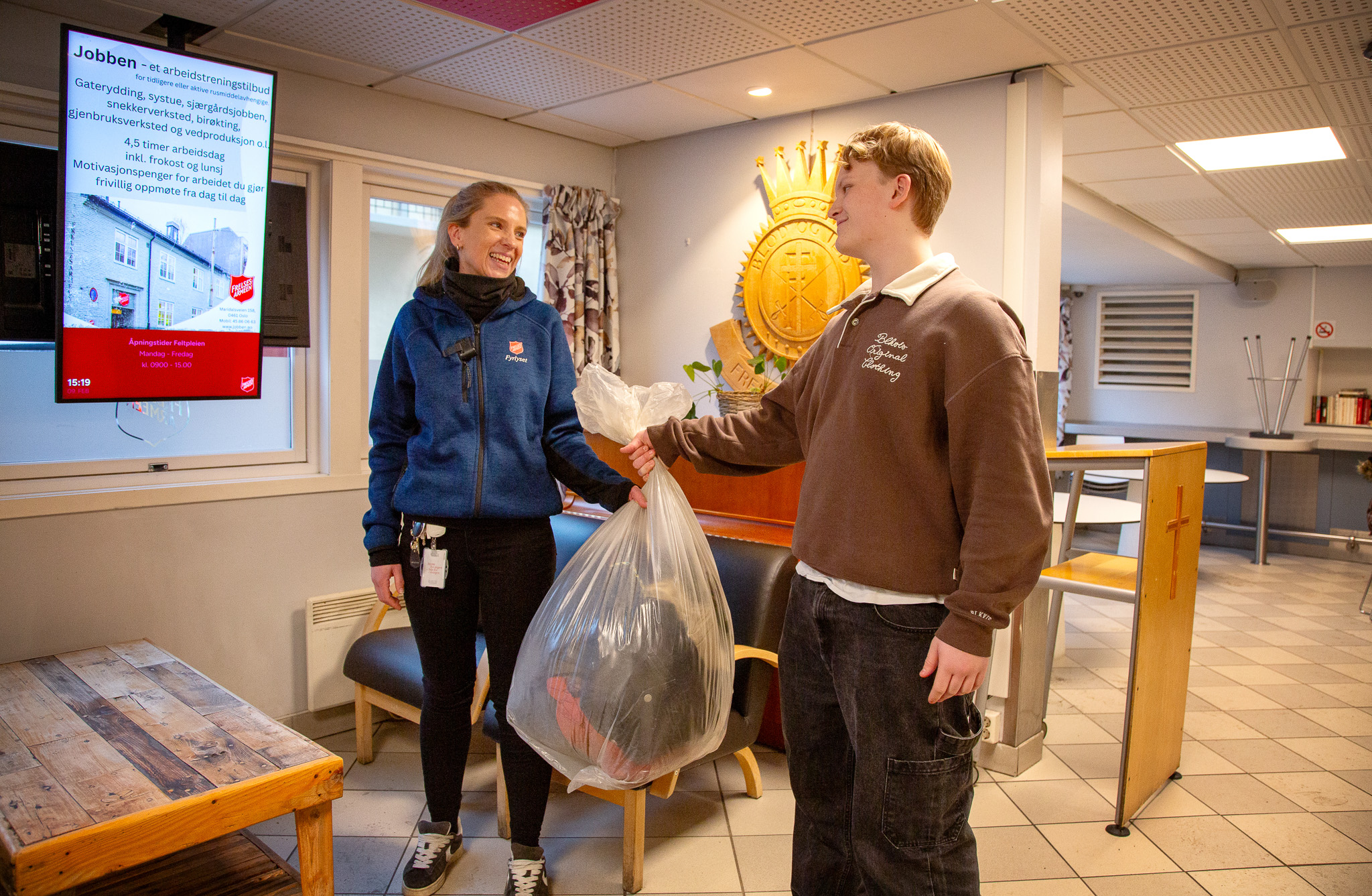 Blond kvinne i blå flisgenser tar imot en pose med klær fra en ung skolegutt inn i et lyst lokale