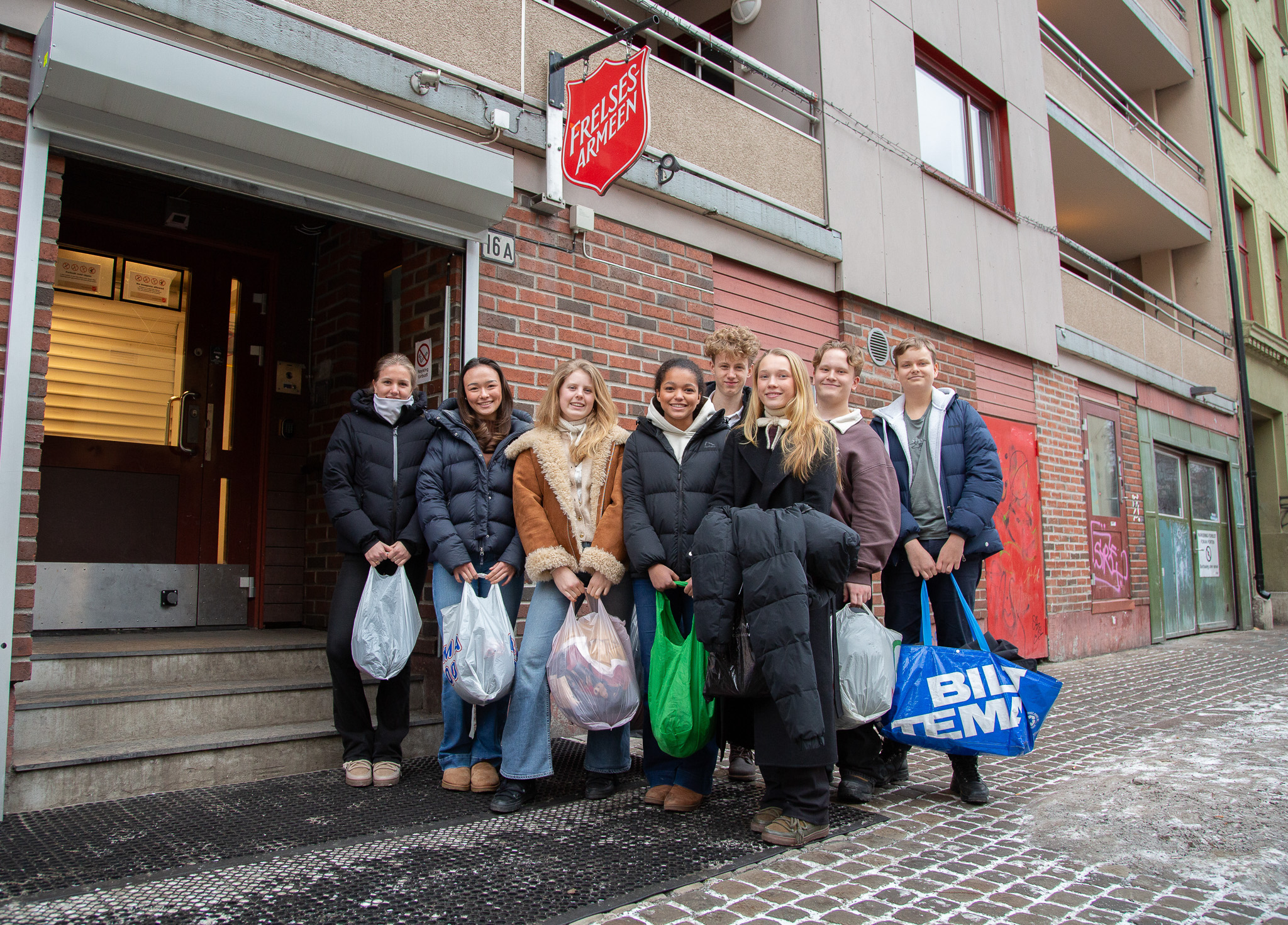 Foto: syv ungdomskoleelever bærer poser, står ute på gaten utenfor et skilt med Frelsesarmeen