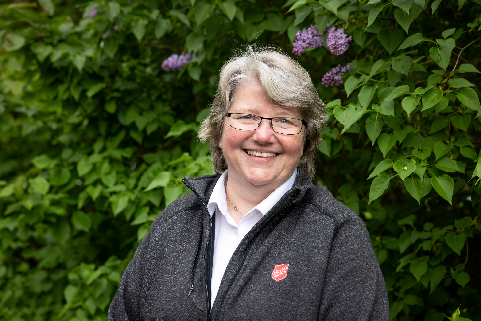 Foto: SMilende kvinne med grått hår og briller i flisjakke med skjold fra Frelsesarmeen.