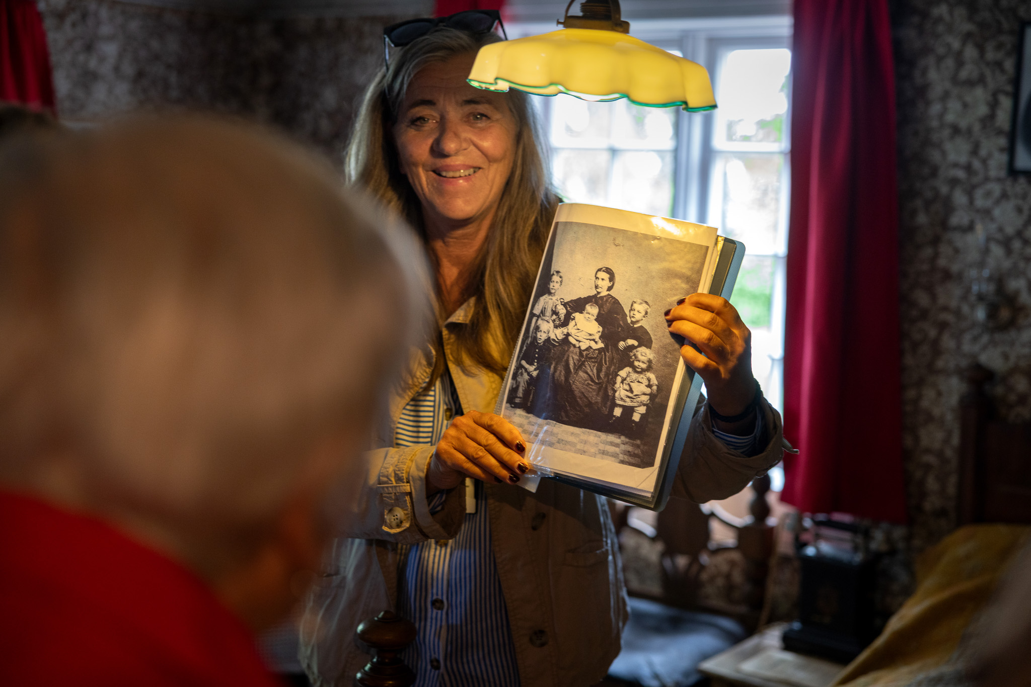 Foto: Smilende kvinne i 60-årene viser fram et svart-hvitt fotografi inne i et gammelt hus.