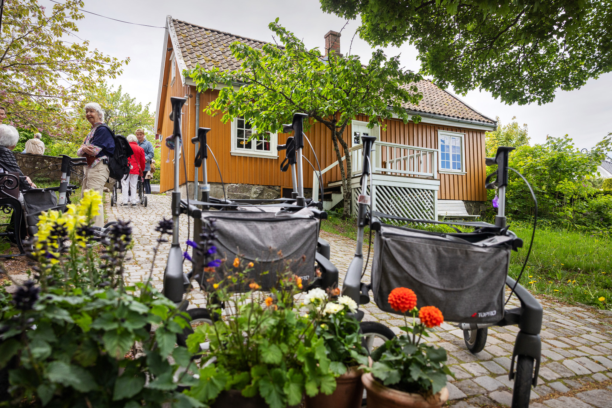 Foto: Guloransje hus i bakgrunnen, eldre mennesker på en vei og et par tullatorer står parkert helt nederst, fra der fotografen tar bildet.