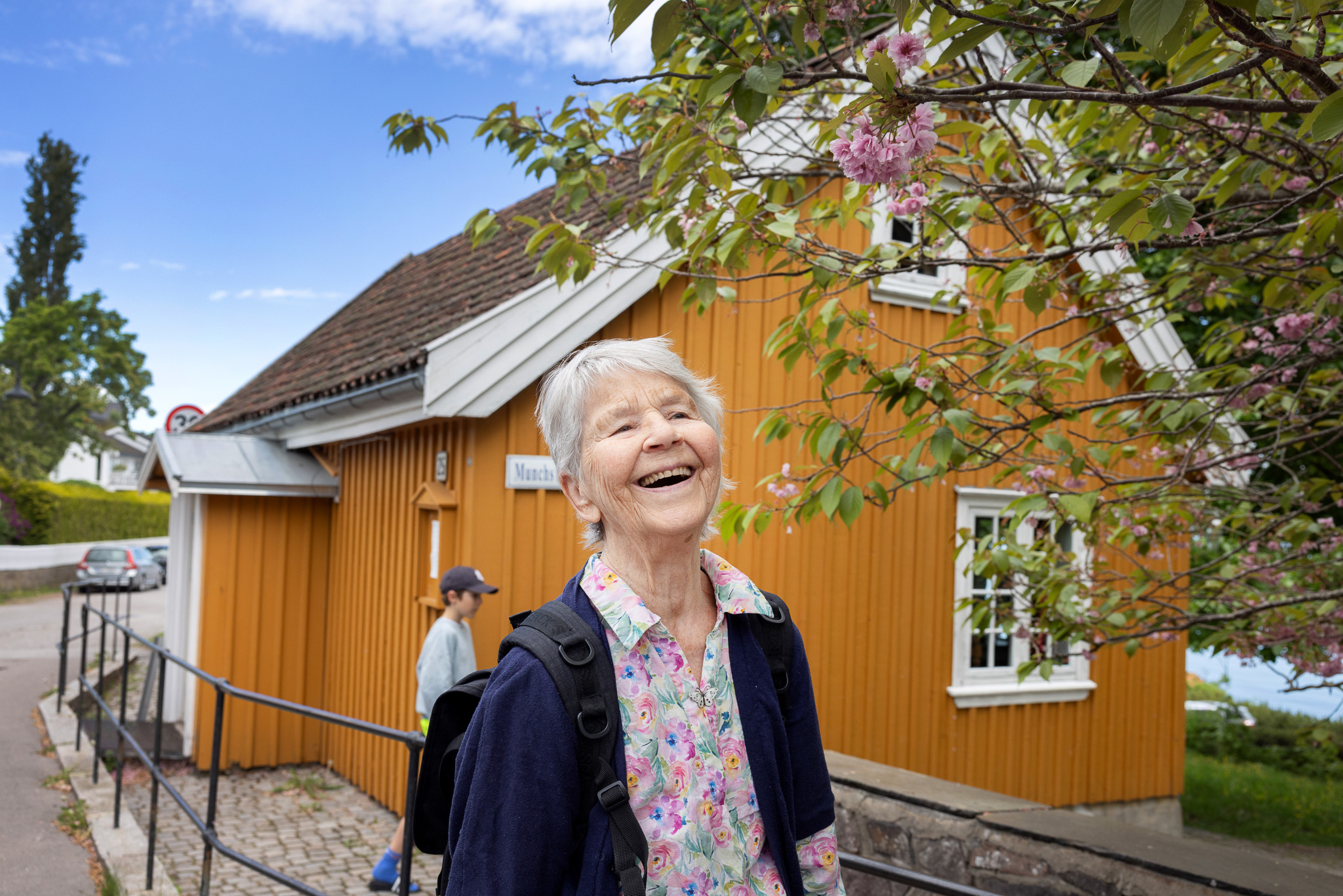 Foto: Kvinne med hvitt hår, smiler foran et lite guloransje hus.