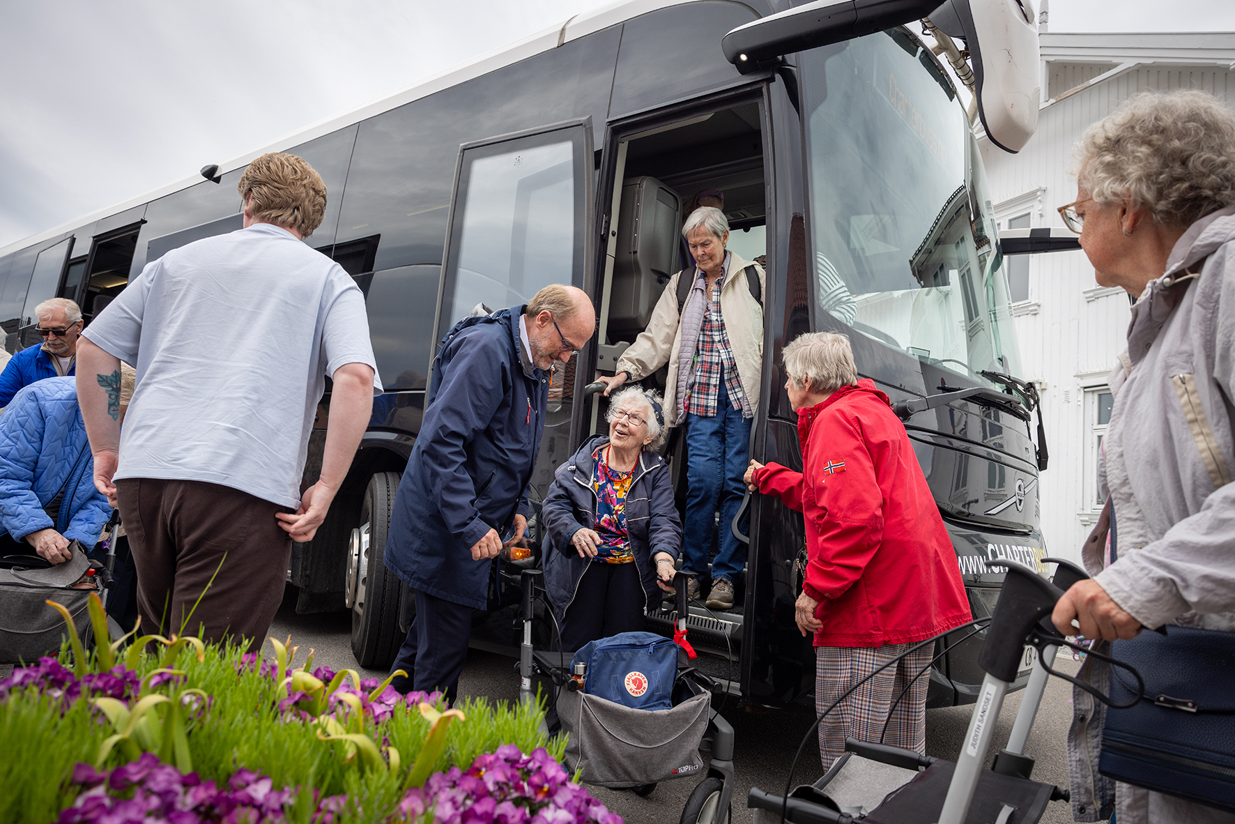 Foto: En gruppe eldre mennesker står utenfor en buss. En kvinne står i trappa opp.