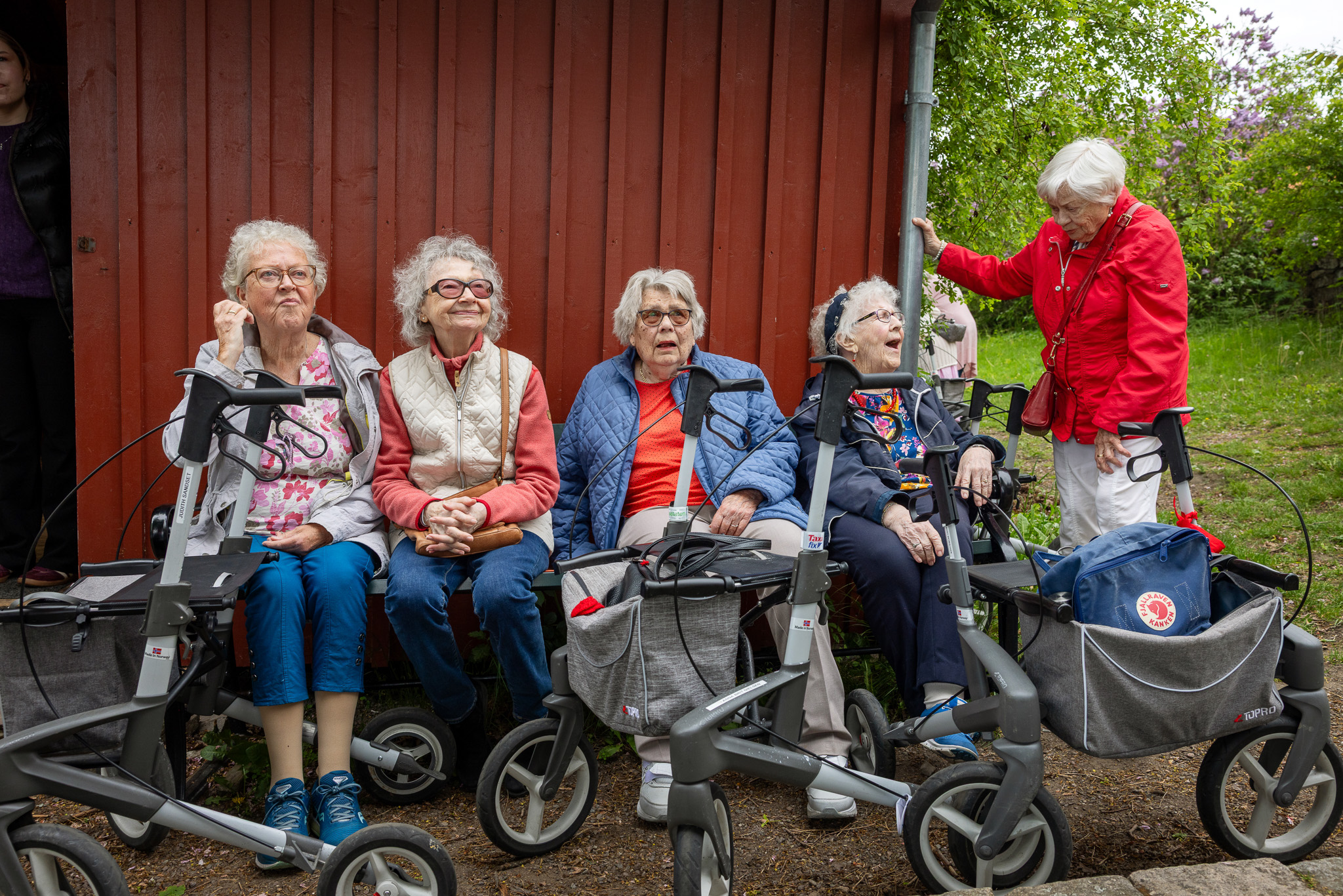 Foto: En gruppe blide, eldre er ute i en hage med grønt gress og trær. Sitter langs en vegg omkranset av rullatorer.