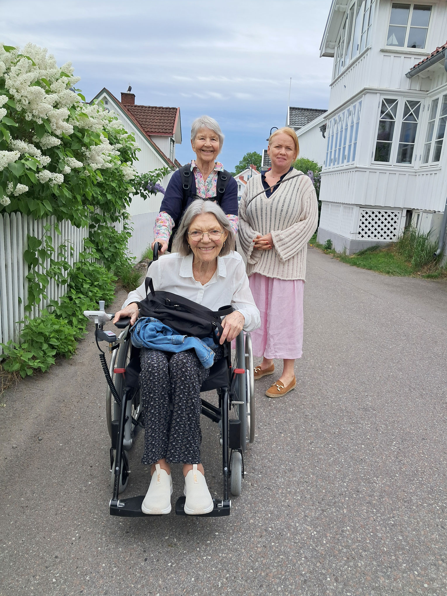 Foto: To damer triller en tredje dame i rullestol nedover en gate med blomstrende, hvite syriner og gamle trehus.