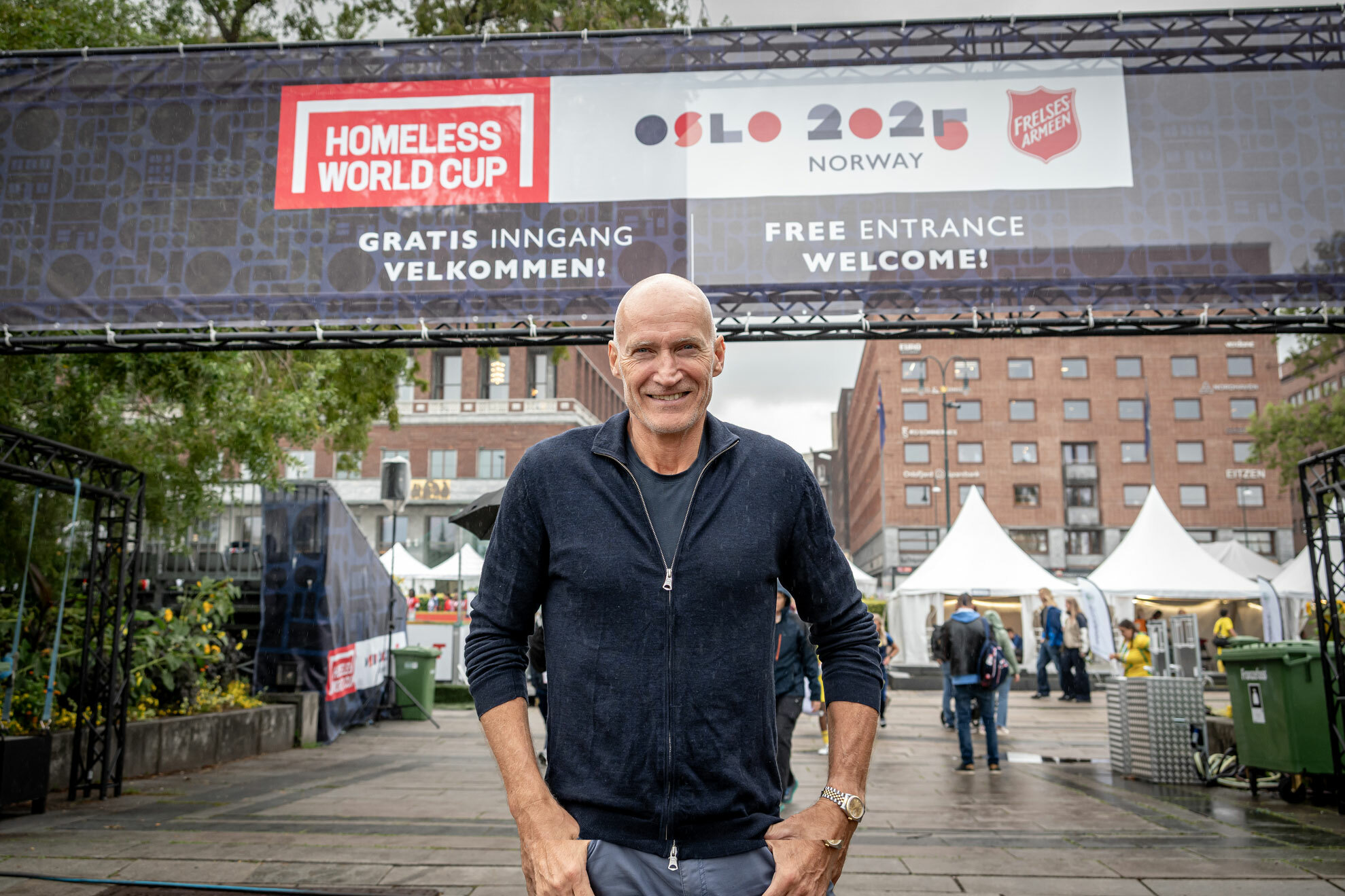Foto: Mann i 60-årene med blank isse står ute på en åpen plass. Bak ser vi rådhuset i Oslo og et banner der det står Homeless World Cup Oslo 2025 Norway og logoen til Frelsesarmeen.