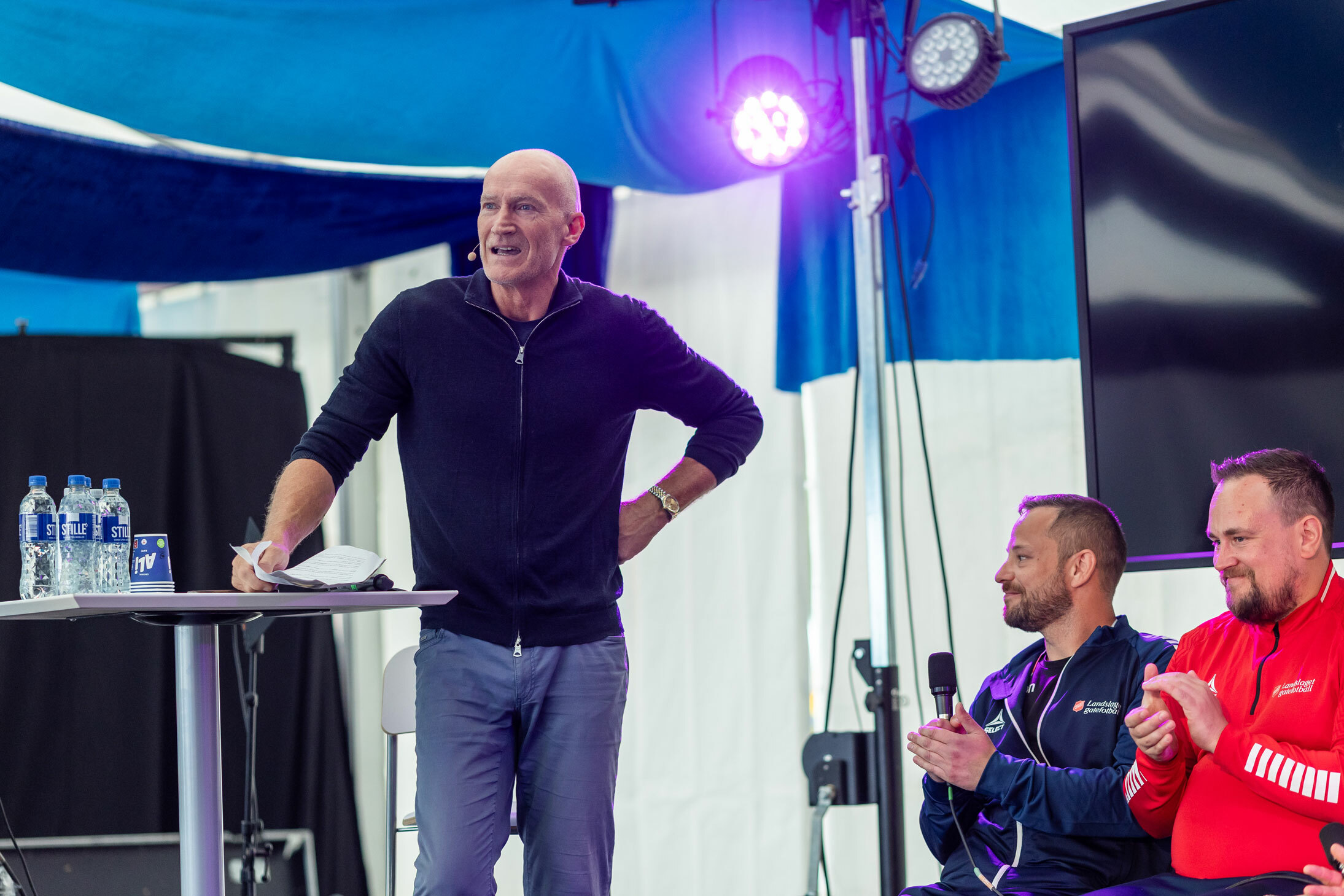 Foto: Mann i 60-årene med blank isse står ved et bord på en scene. Bak ham sitter noen menn i treningstøy på rekke.