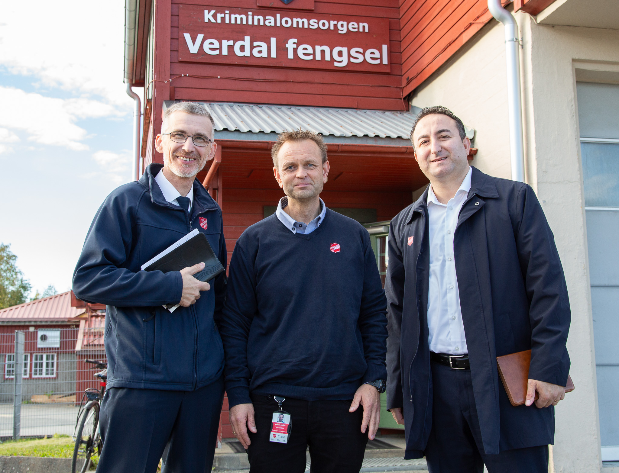 Foto: Tre menn kledd i lette ytterjakker. Står ved siden av hverandre og smiler. I bakgrunnen er er rødt bygg.